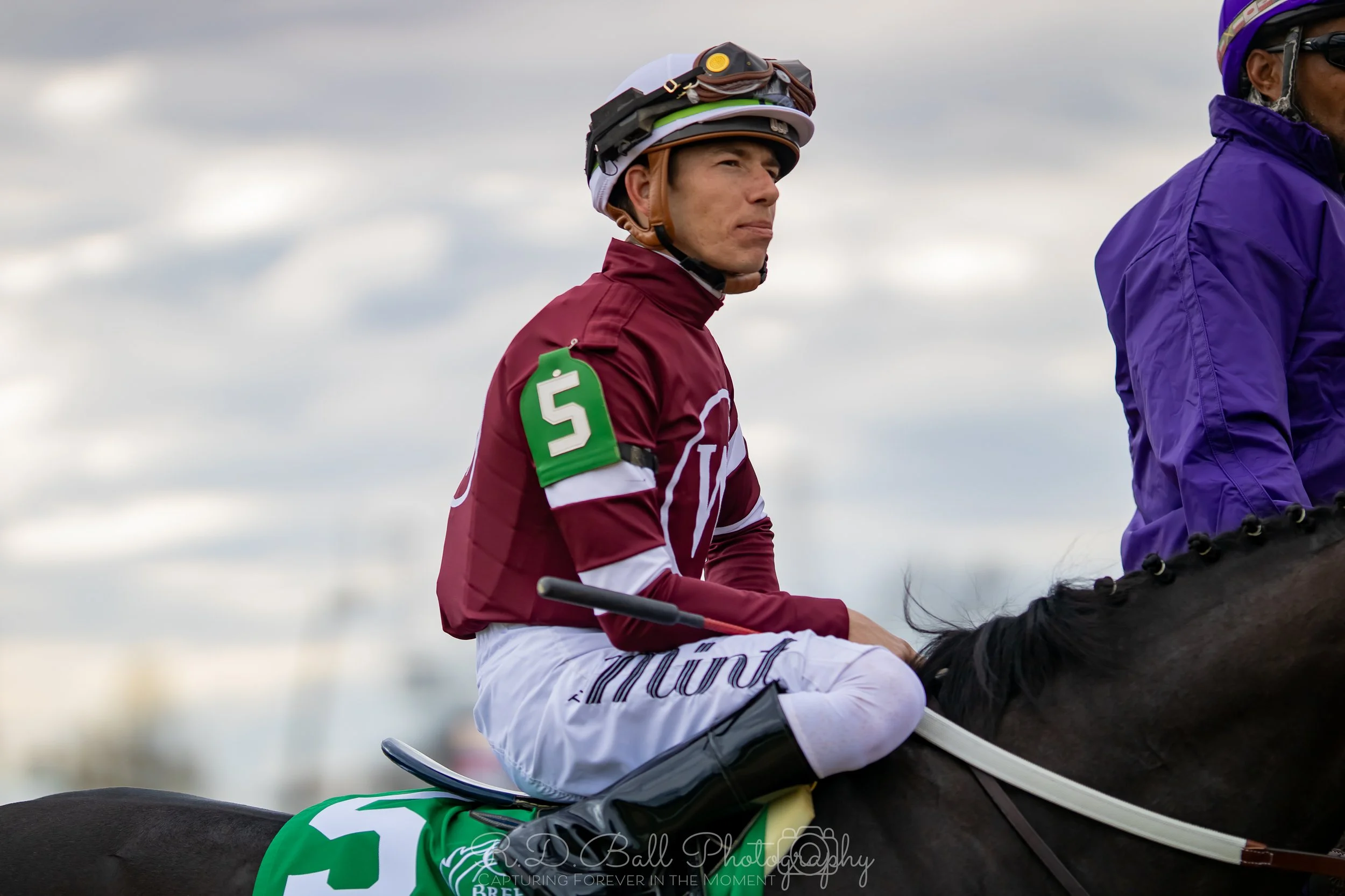 A jockey dressed in maroon and white riding gear, sitting on a dark horse, during an outdoor horse racing event. The jockey is wearing a helmet with goggles, and there is another rider in purple clothing beside him.