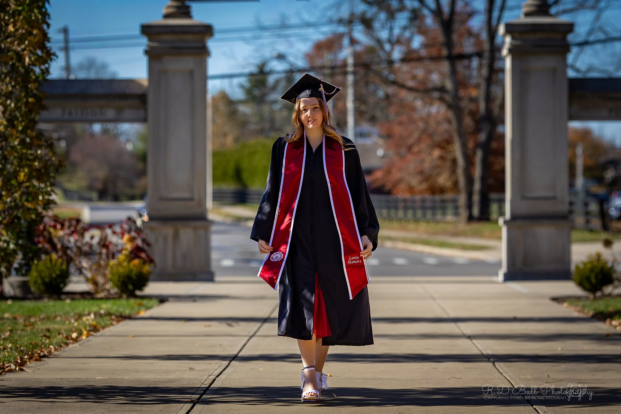 A young woman in a graduation cap and gown walking outdoors on a sidewalk on a sunny day, with trees and a stone archway in the background.