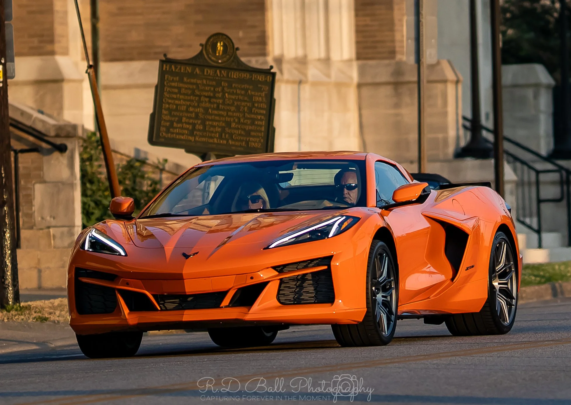 An orange Chevrolet Corvette sports car with sleek design driving on a city street during the day, with a historical building and a plaque in the background.