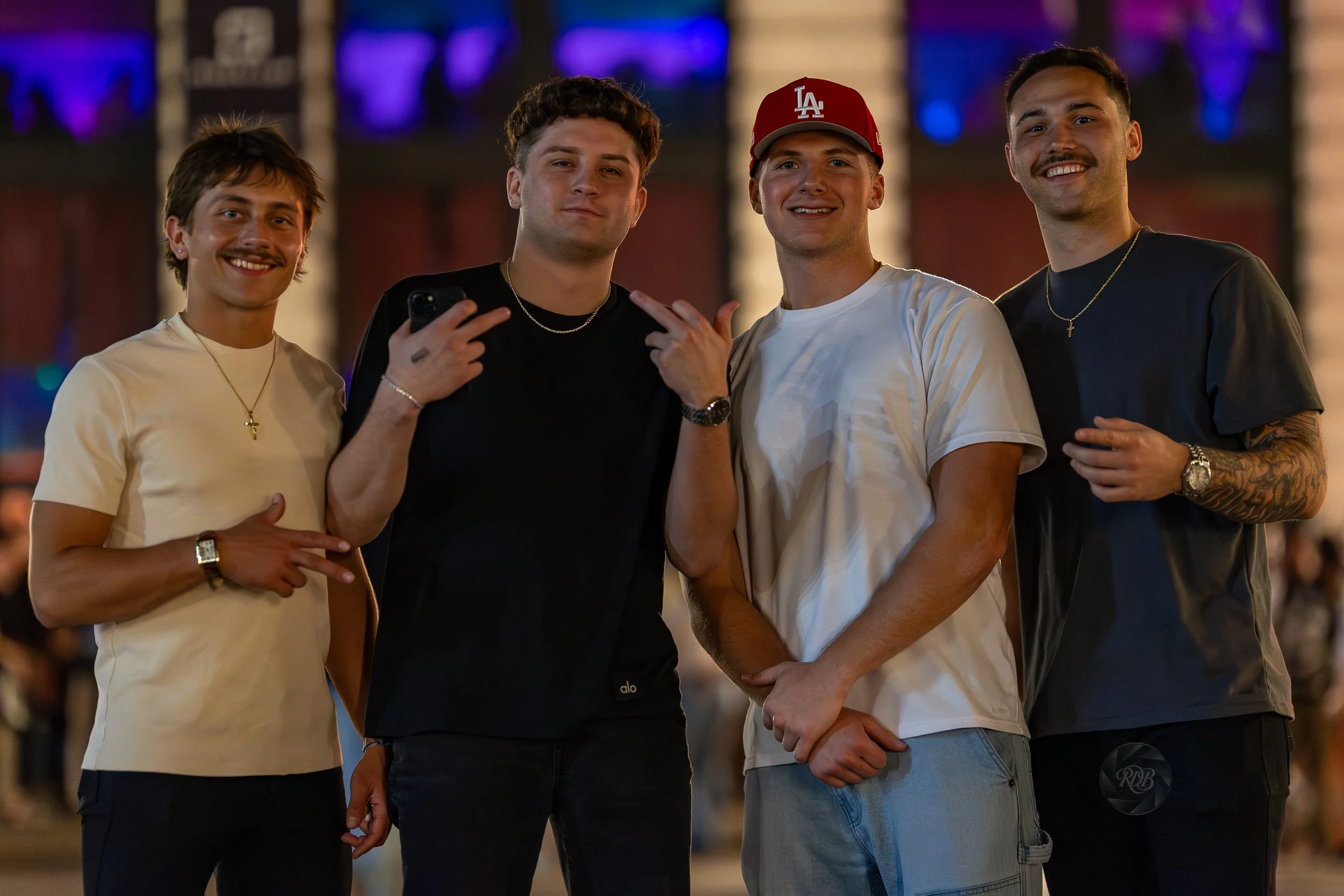 Four young men posing together at night, smiling with a blurred crowd and colorful lights in the background.