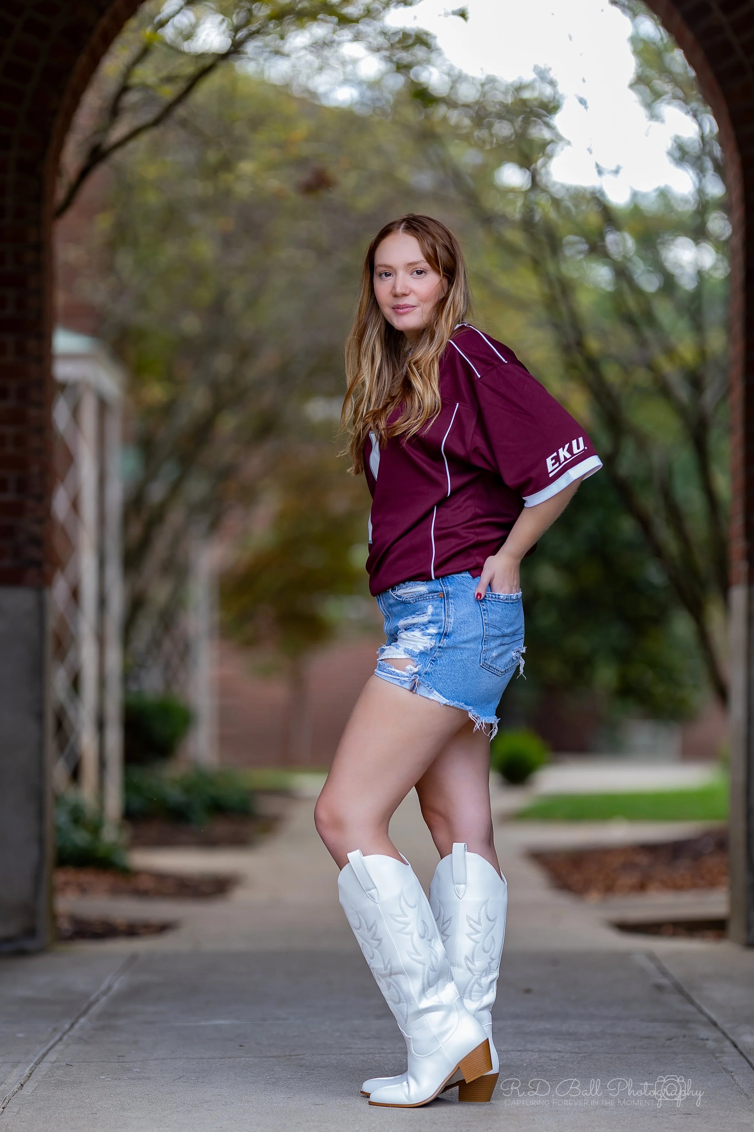A young woman with long wavy hair wearing a maroon sports jersey, denim shorts, and white cowboy boots stands outdoors under a brick archway, with trees in the background.