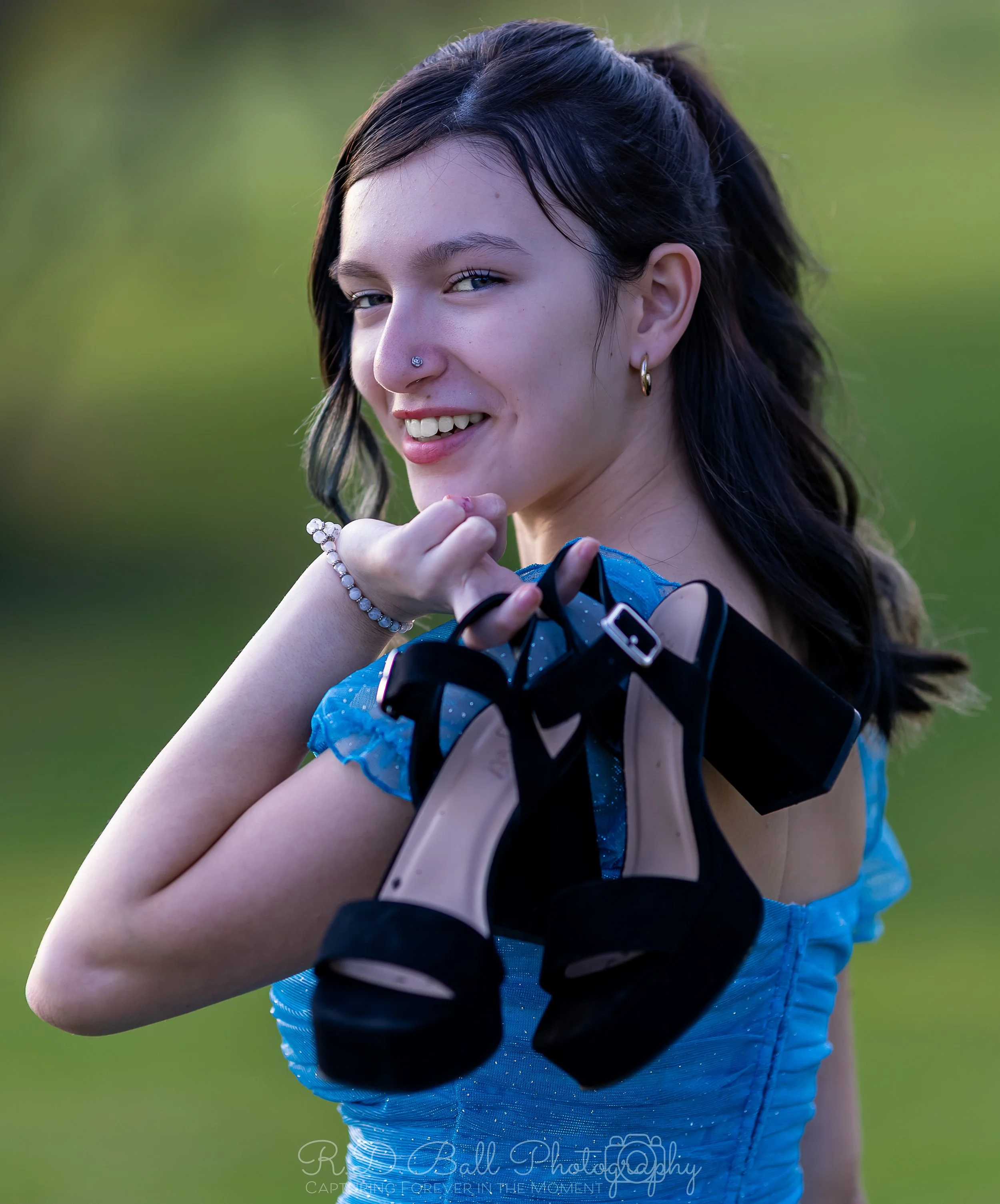 Young woman with dark hair smiling, holding black high-heeled sandals and looking over her shoulder outdoors.