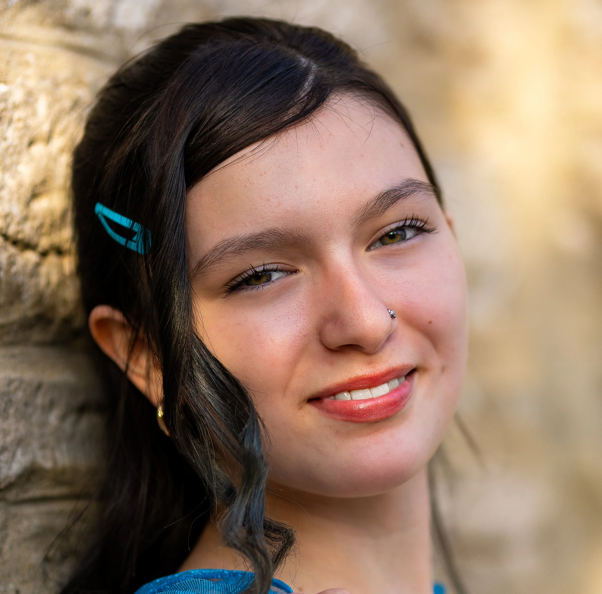 Close-up of a young woman with dark hair, a blue hair clip, a nose ring, and a bright smile, leaning against a stone wall.