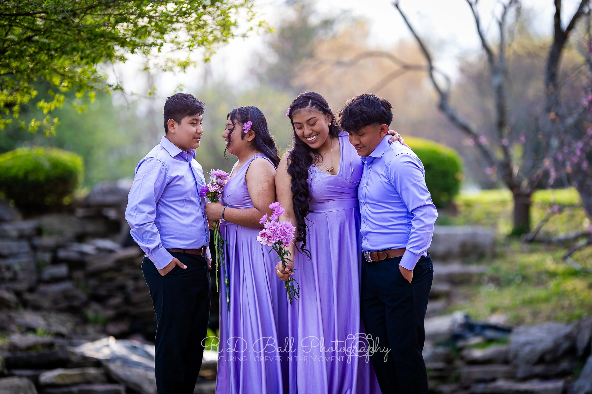 Four young people, two boys and two girls, wearing matching purple dresses and shirts, standing outdoors in a garden, holding flowers, smiling, and embracing each other in a joyful moment.
