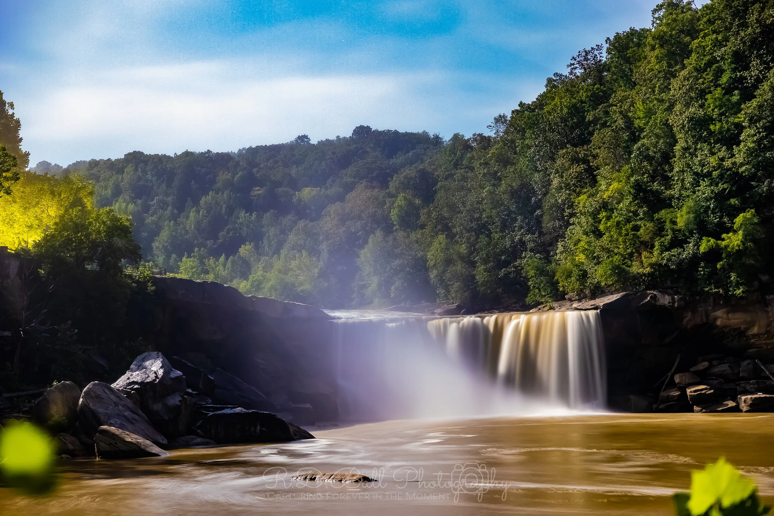 A waterfall flowing into a river surrounded by lush green trees and a blue sky.