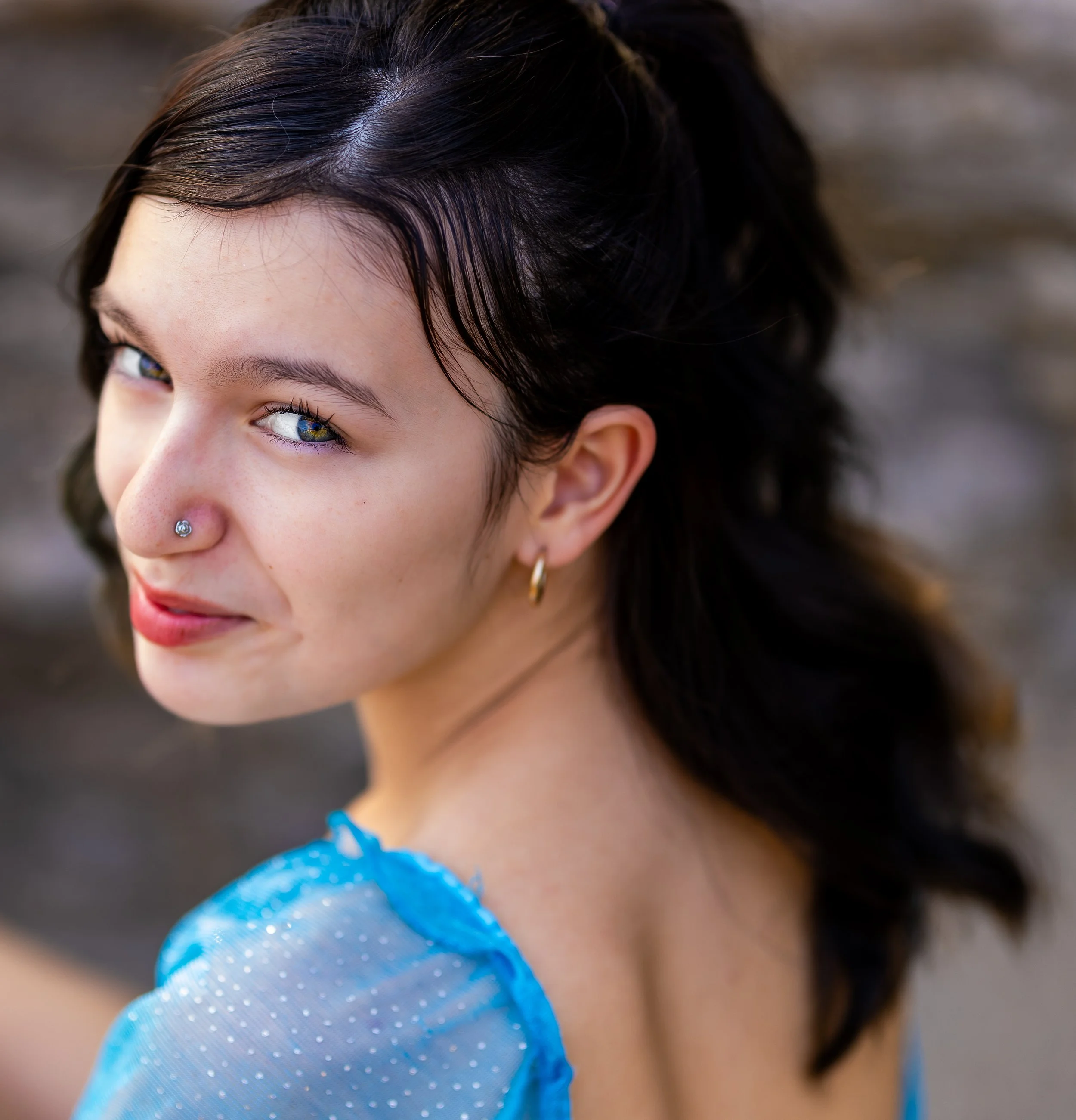 Close-up of a young woman with dark hair, a nose piercing, and an earring, wearing a blue sheer polka dot top, looking over her shoulder with a slight smile.