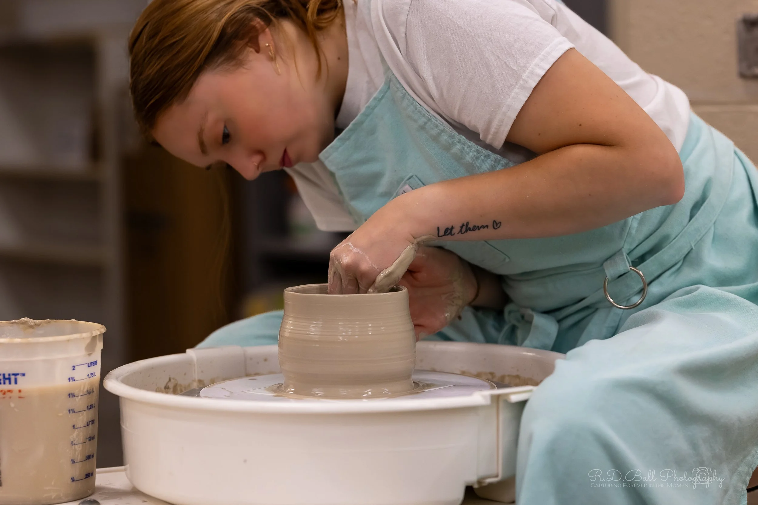A woman with short red hair and a tattoo reading 'Let them' on her forearm is working on a pottery wheel, shaping a clay pot.