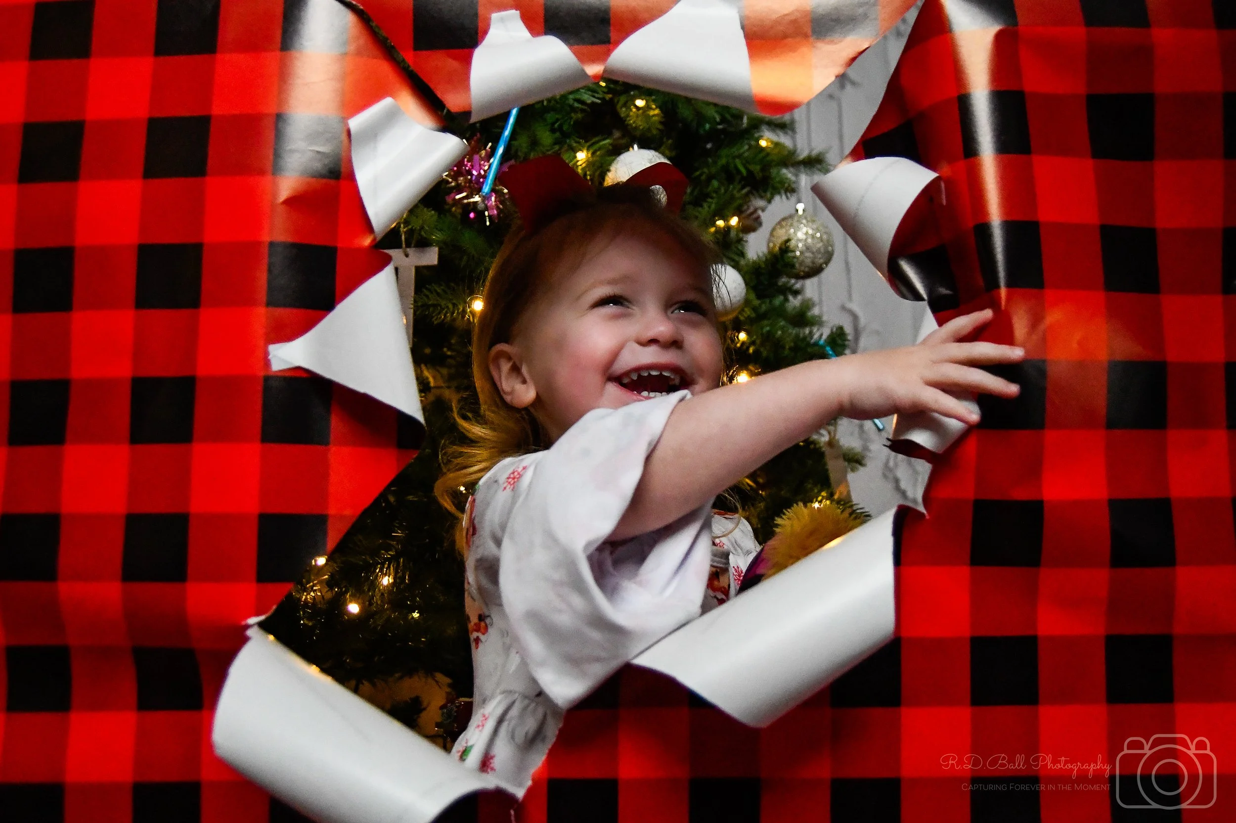 Child peering through a torn hole in a large, red and black checkered wrapping paper, with a decorated Christmas tree with lights and ornaments in the background.