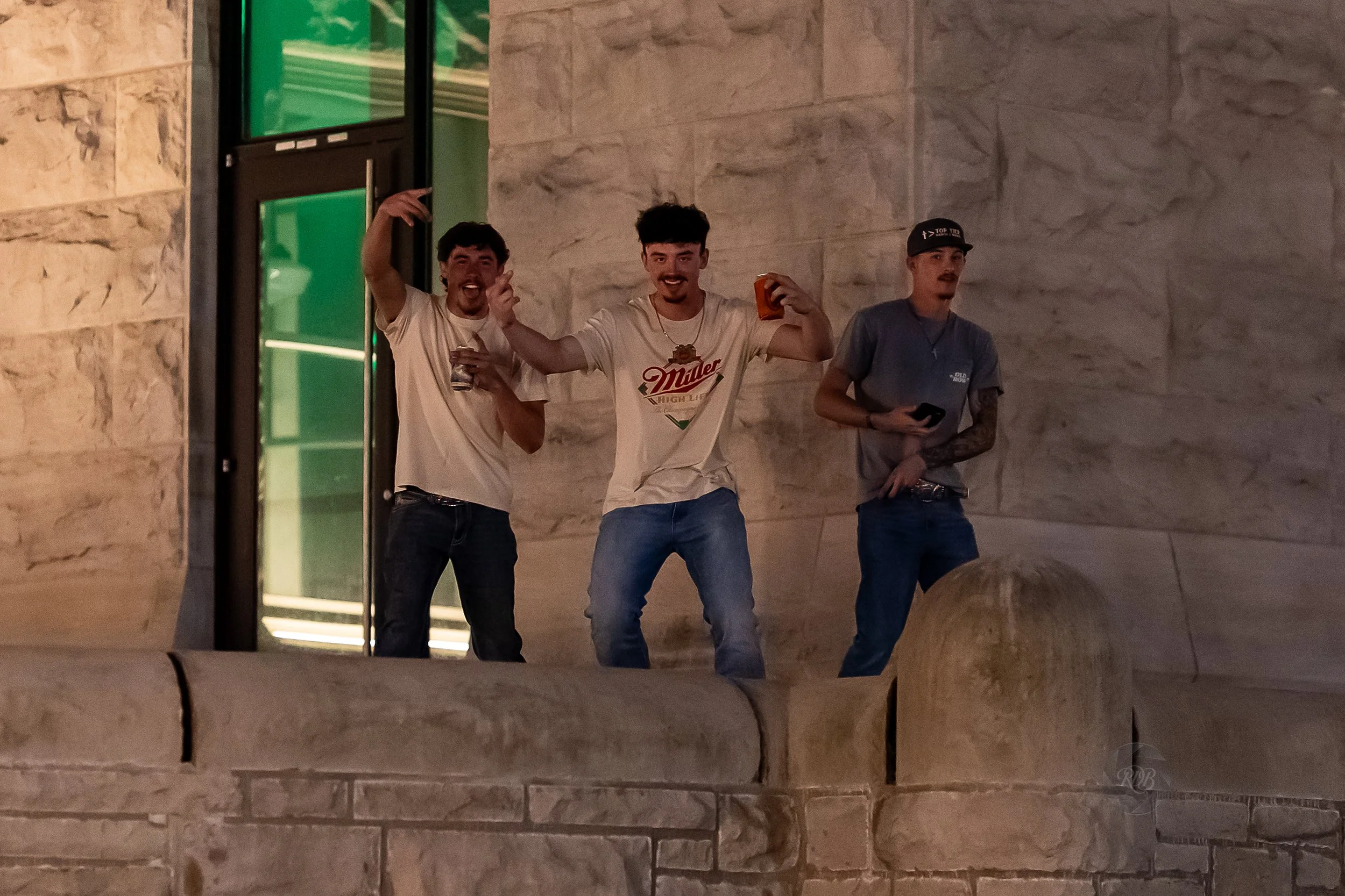 Three young men standing near a brick wall, some holding drinks and one with his phone, smiling and posing for the photo at night.