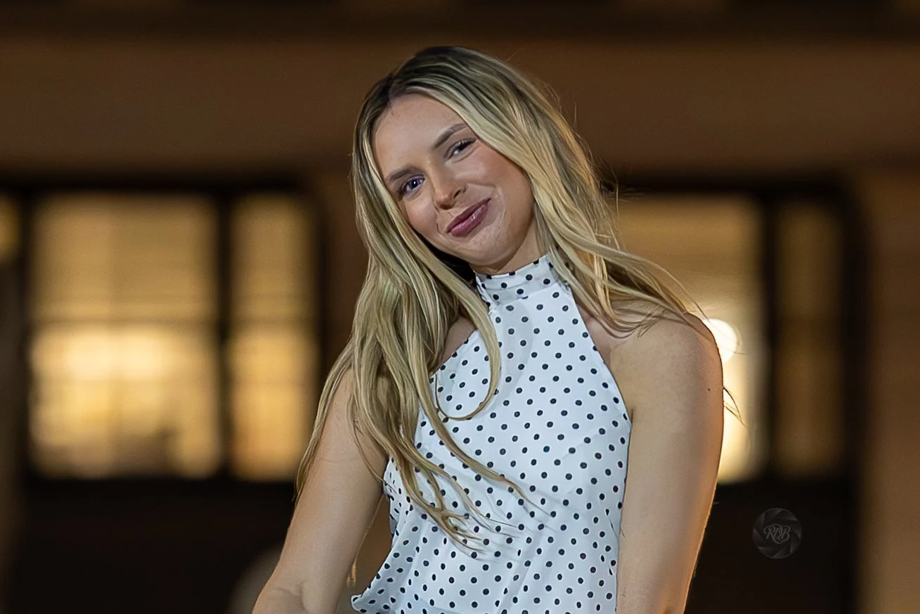A young woman with long blonde hair wearing a sleeveless white top with black polka dots, smiling at the camera against a blurred indoor background.