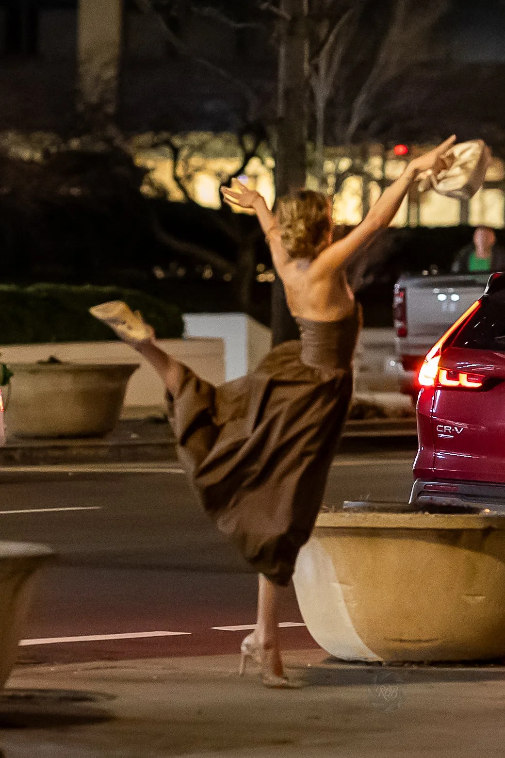 A woman in a strapless brown dress is dancing or jumping on a sidewalk at night, with one foot on a concrete planter and her arms raised, holding a bag. There is a red car nearby and blurred background trees and streetlights.
