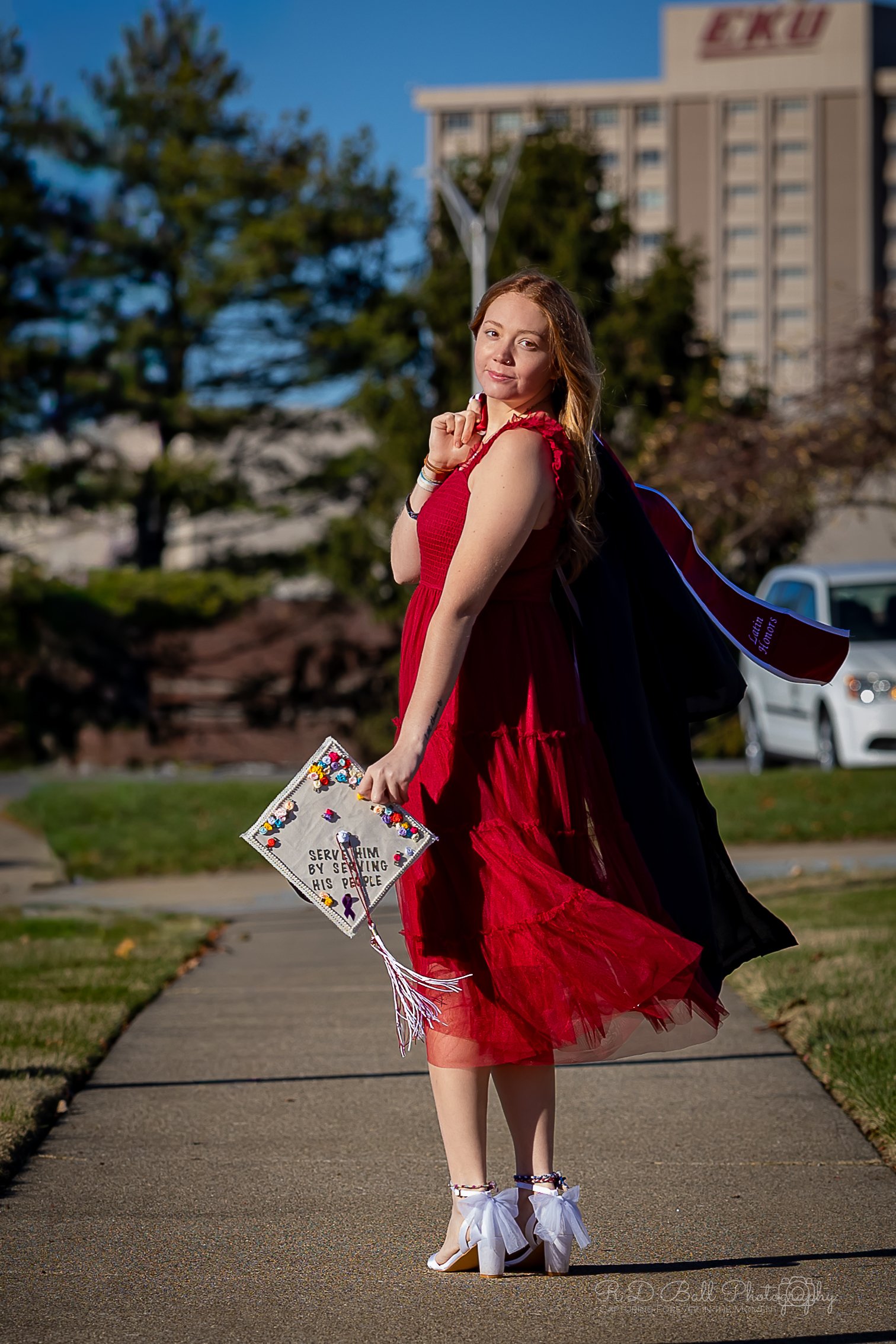 A young woman in a red dress holding a decorated graduation cap with a quote, dressed in white shoes with bows, standing outdoors on a sidewalk with trees and buildings in the background.