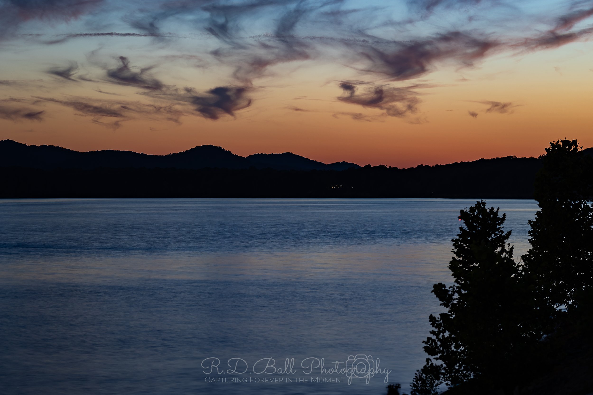 A serene lake at sunset with dark silhouettes of trees in the foreground, calm water reflecting the colorful sky, and mountains in the distance under a partly cloudy sky.