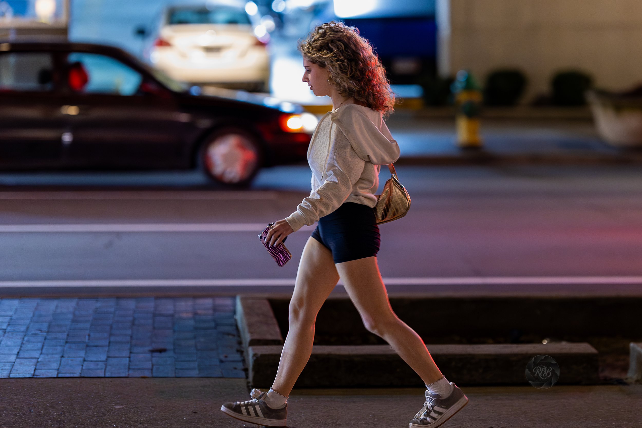 A young woman walking on a city street at night, wearing a light-colored hoodie, black shorts, and Adidas sneakers, carrying a patterned phone case in her hand and a small bag on her shoulder, with cars and blurred city lights in the background.