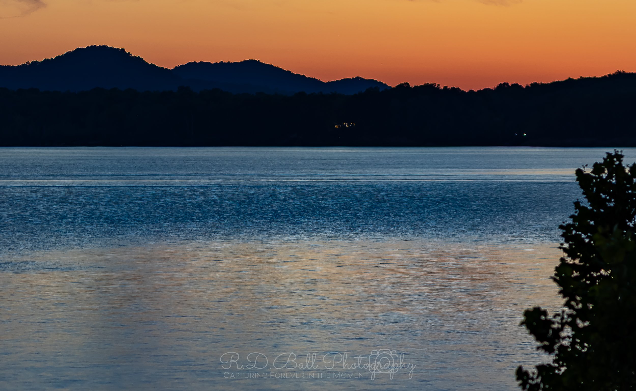 Sunset over a calm lake with silhouetted mountains in the background and a tree branch on the right side.