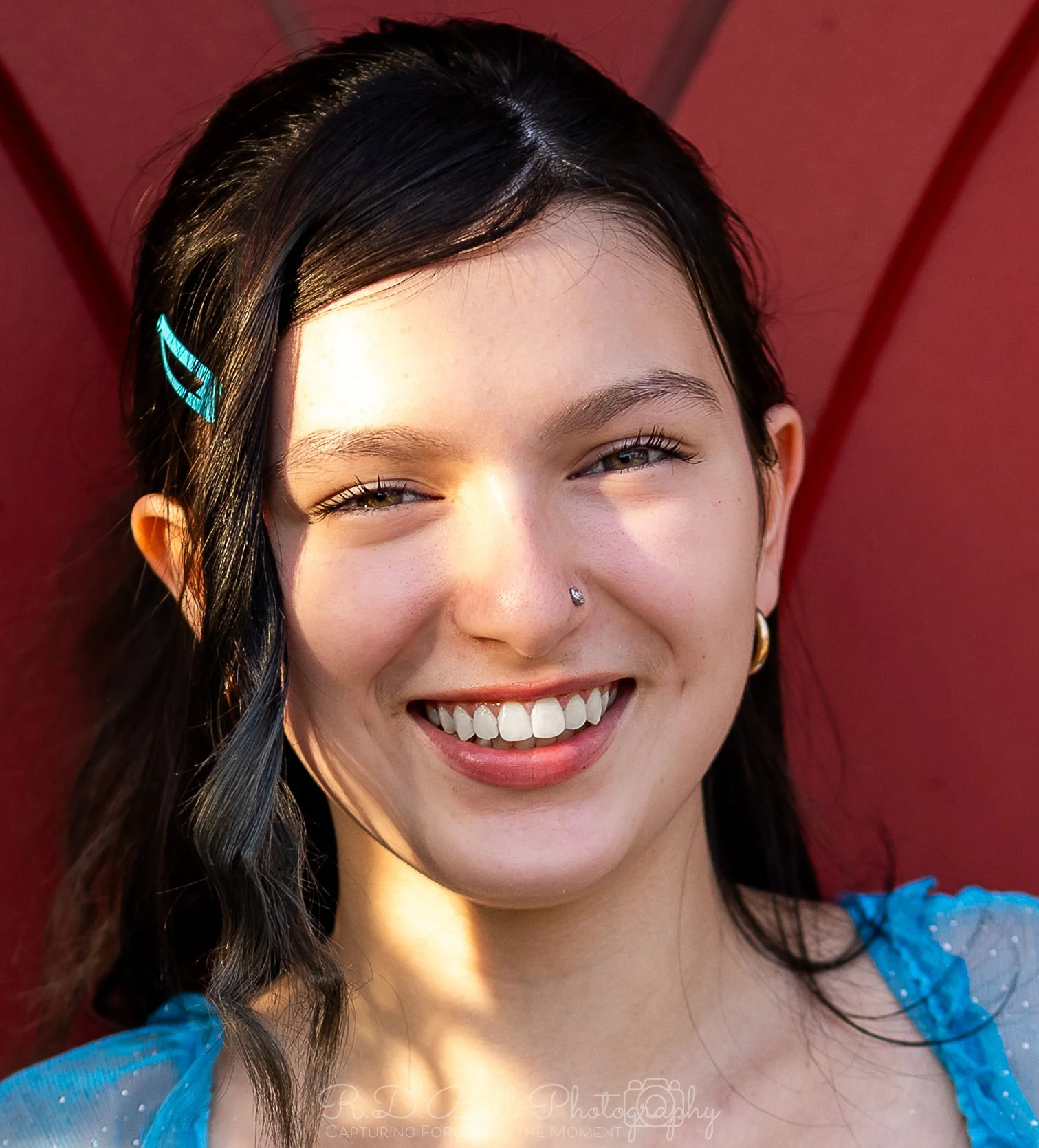 Close-up of a young woman with black hair, blue hair clip, hoop earrings, a nose piercing, and a bright smile, wearing a blue top, with a red background.