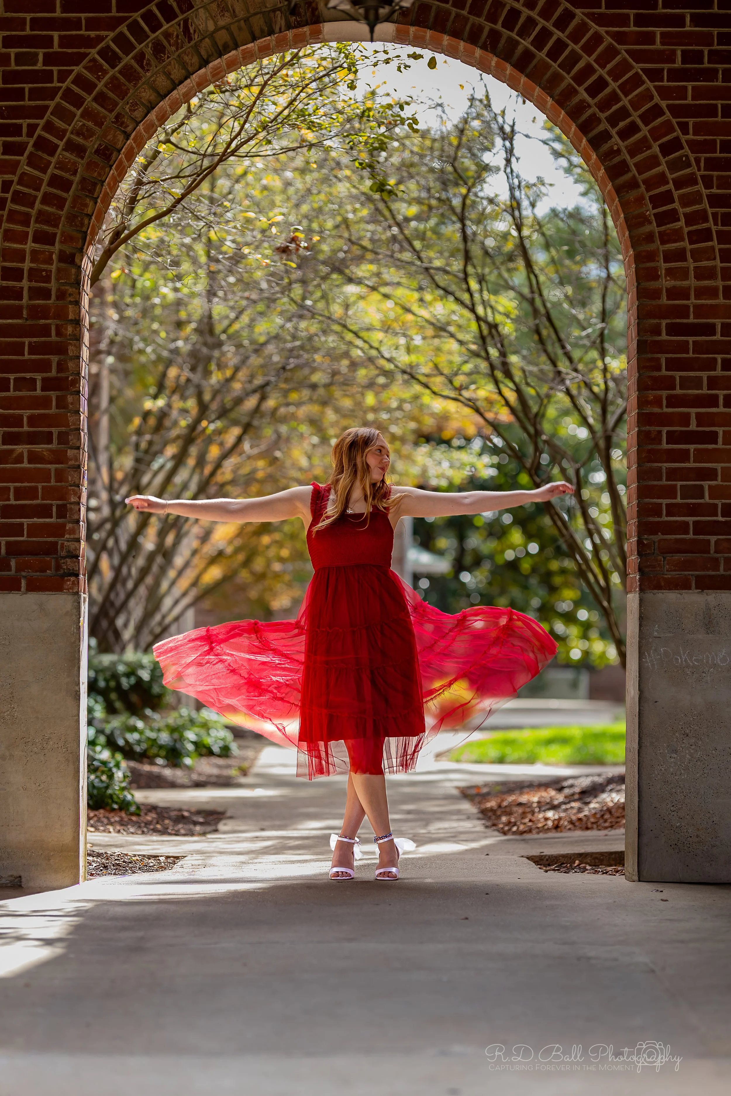 A woman in a red dress twirling under an archway at a park during fall, with trees in the background.