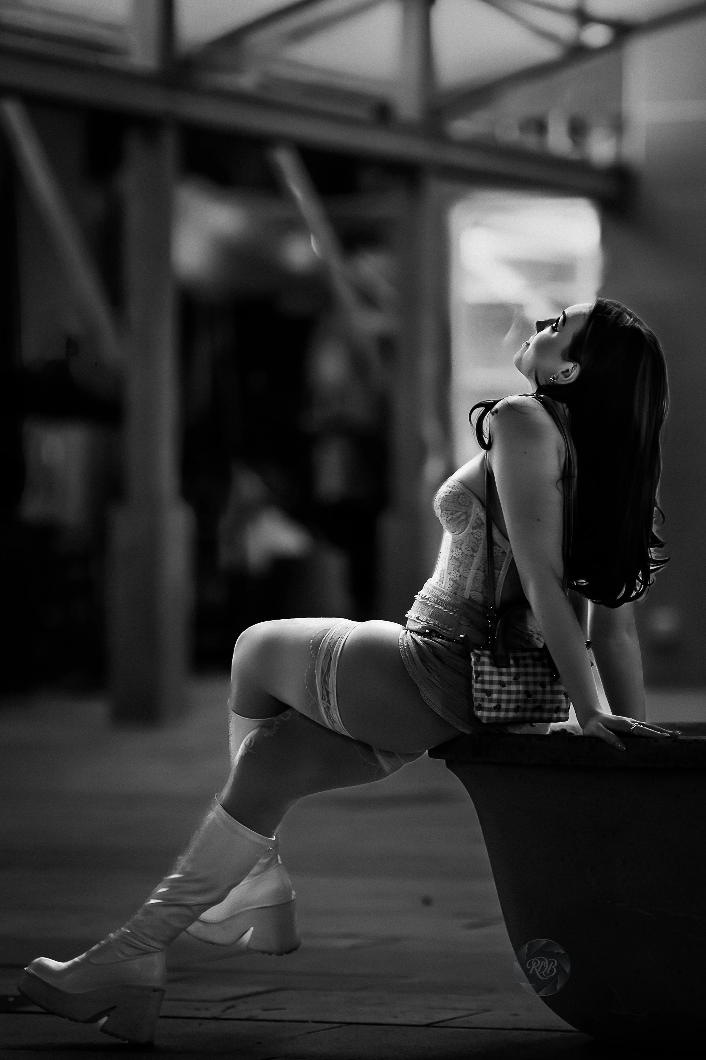 A woman sitting on a bench, wearing thigh-high boots, denim shorts, and a lace top, with her head tilted back and eyes closed, in a black-and-white photograph.