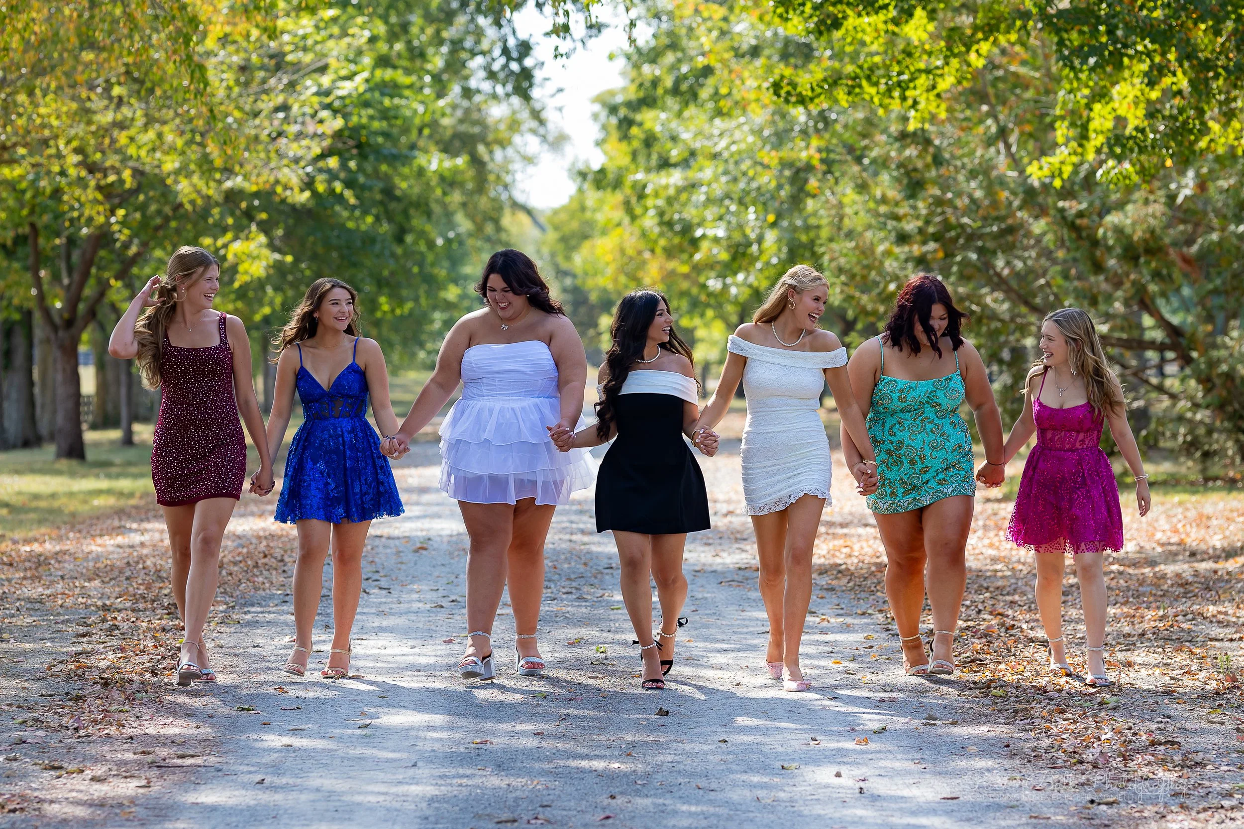 A group of women in colorful dresses holding hands and walking together on a tree-lined path in a park during daytime.