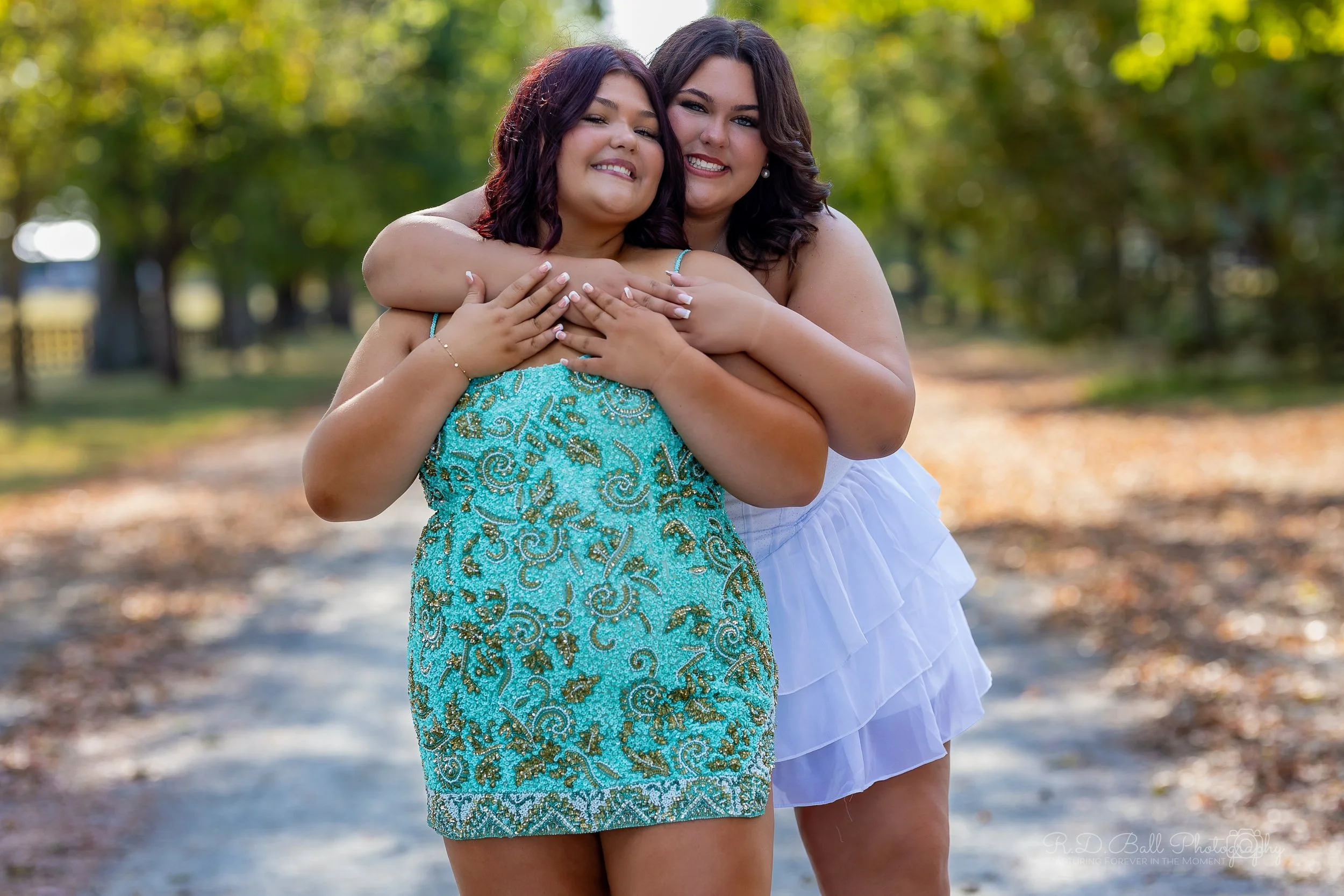 Two women hugging and smiling outdoors on a sunny day, with trees and a dirt path in the background.