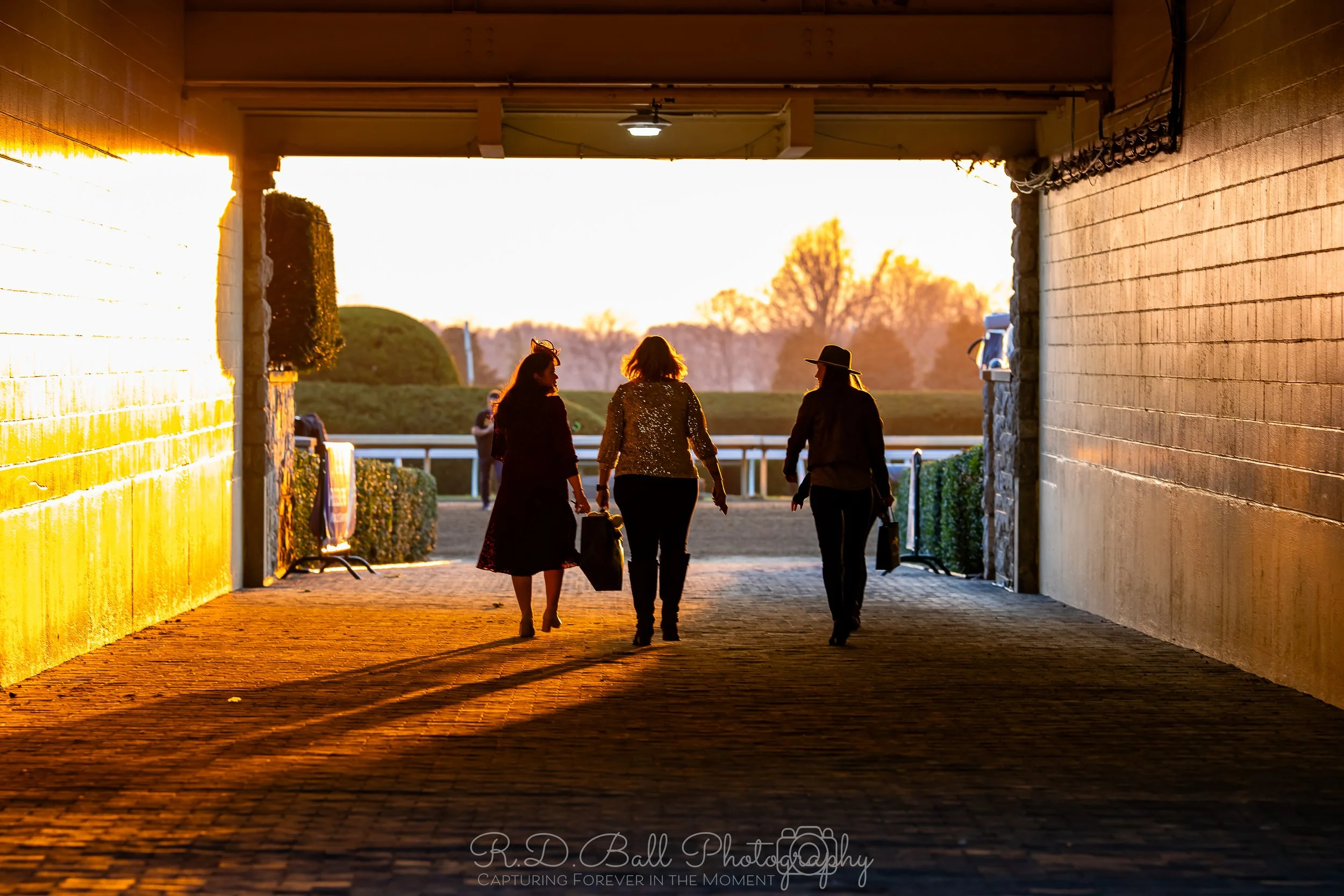 Three women walking through a tunnel at a racetrack during sunset, carrying bags, with a horse racing track visible outside the tunnel.