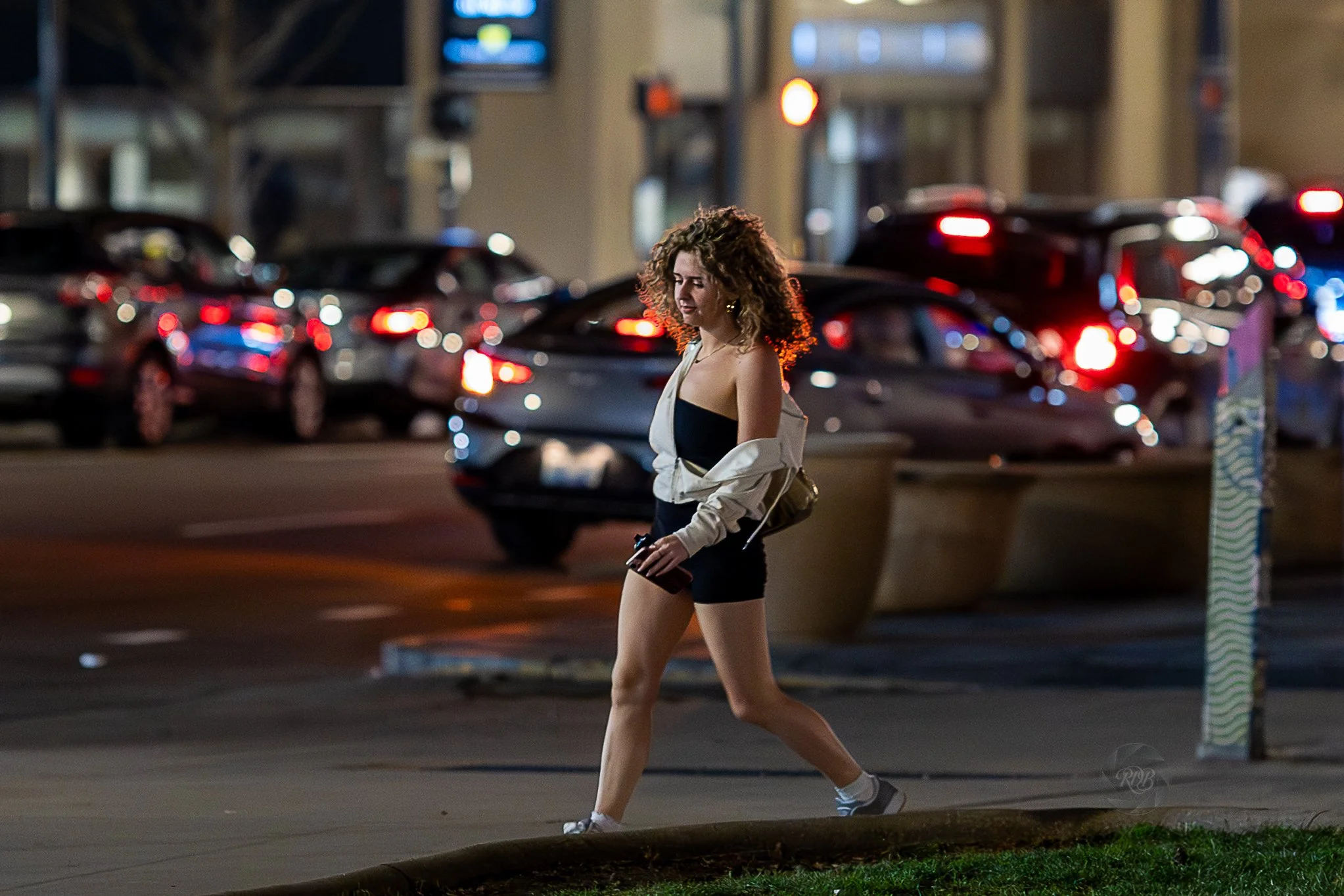 A woman with curly hair walking on the sidewalk at night, wearing a black dress, a light jacket hanging off her shoulders, and sneakers, with city lights and parked cars in the background.