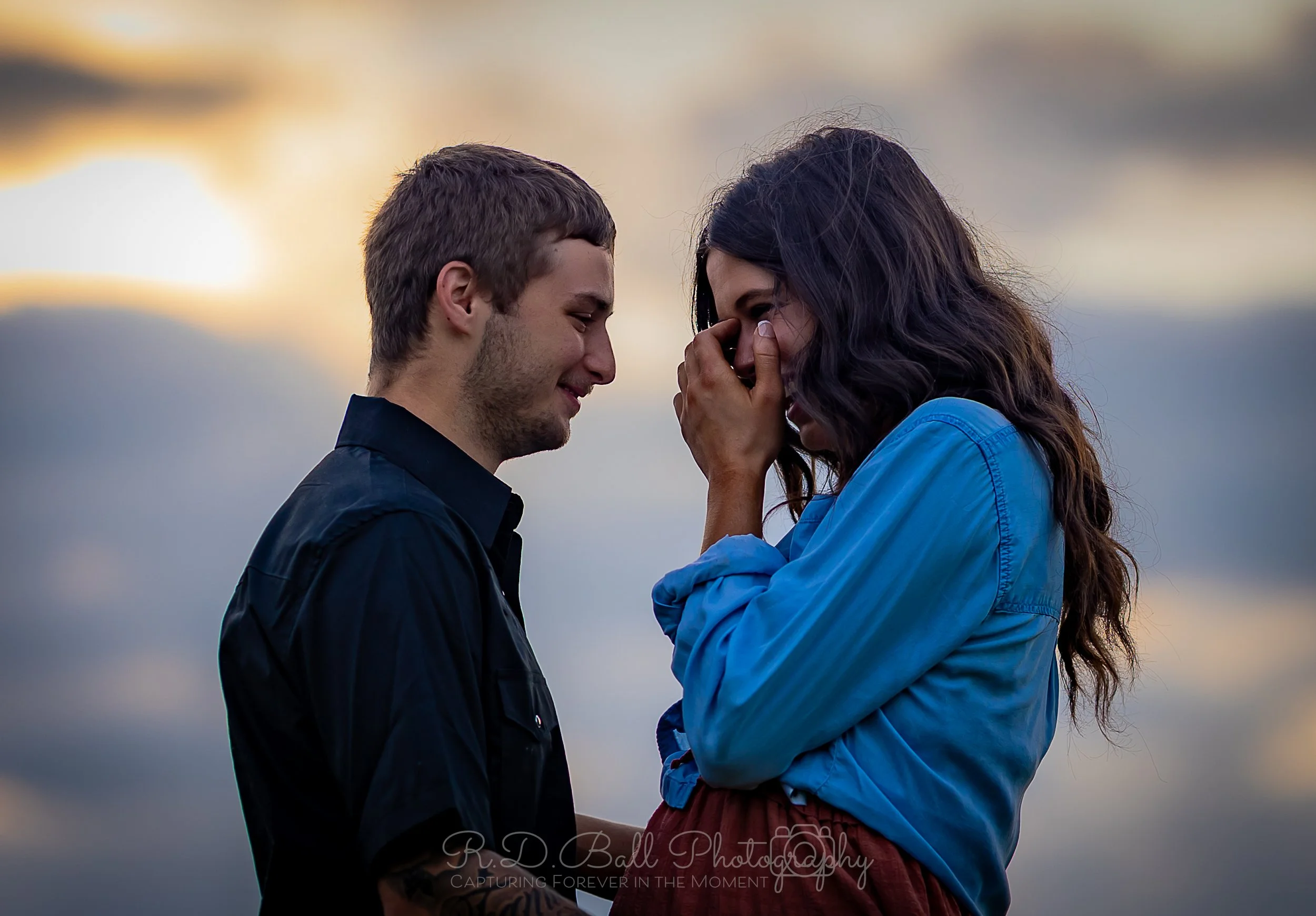 A couple stands close together at sunset on a beach, with the woman holding her hand to her face as she smiles, and the man looking fondly at her.