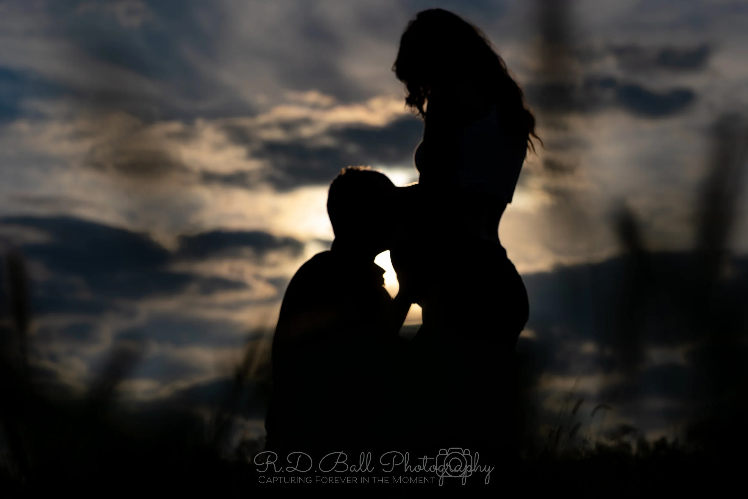 Silhouette of a couple embracing outdoors during sunset with cloudy sky in the background.