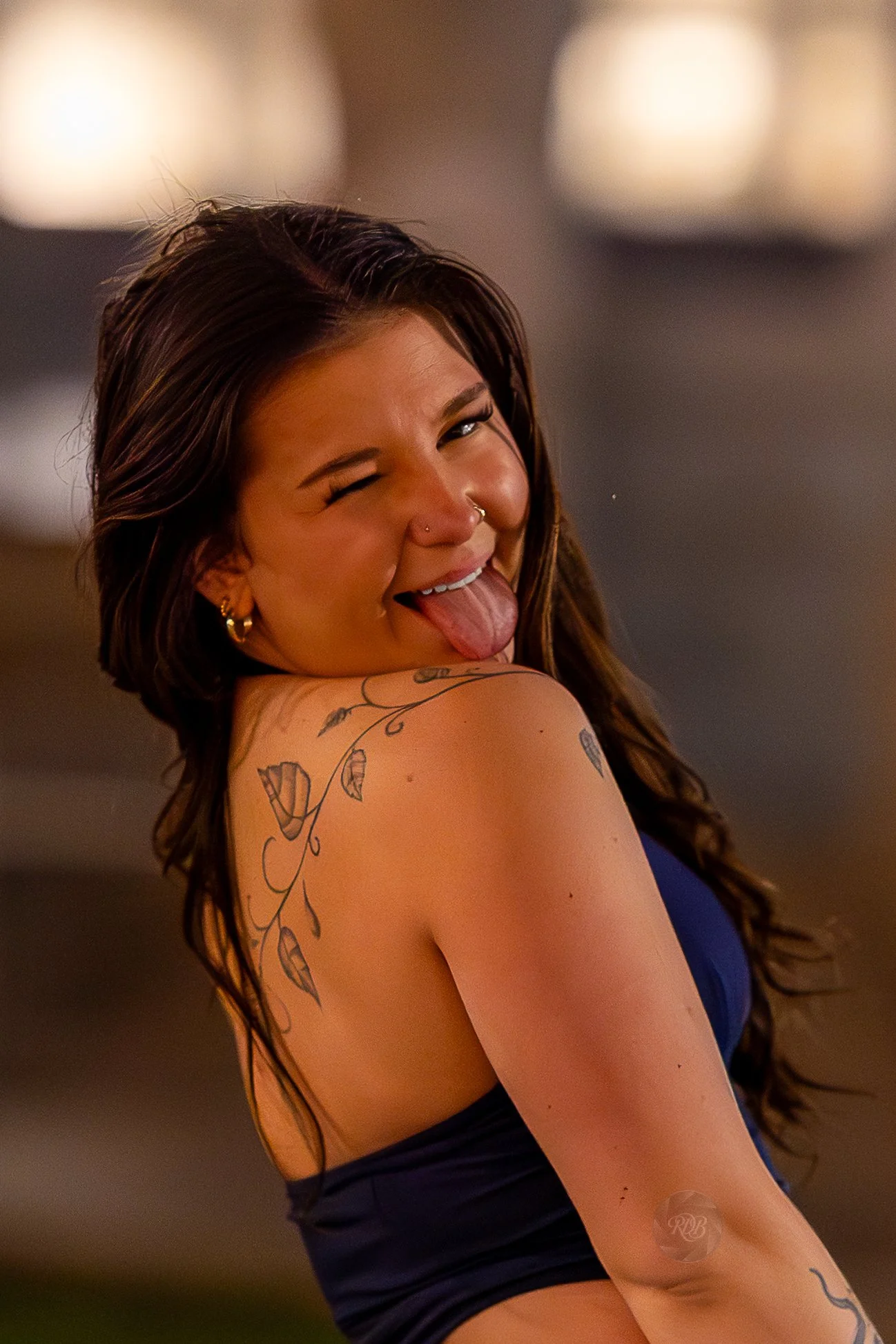 A young woman with dark, wavy hair and tattoos on her shoulder and arm, sticking out her tongue and winking at the camera, wearing a dark top and gold earrings.