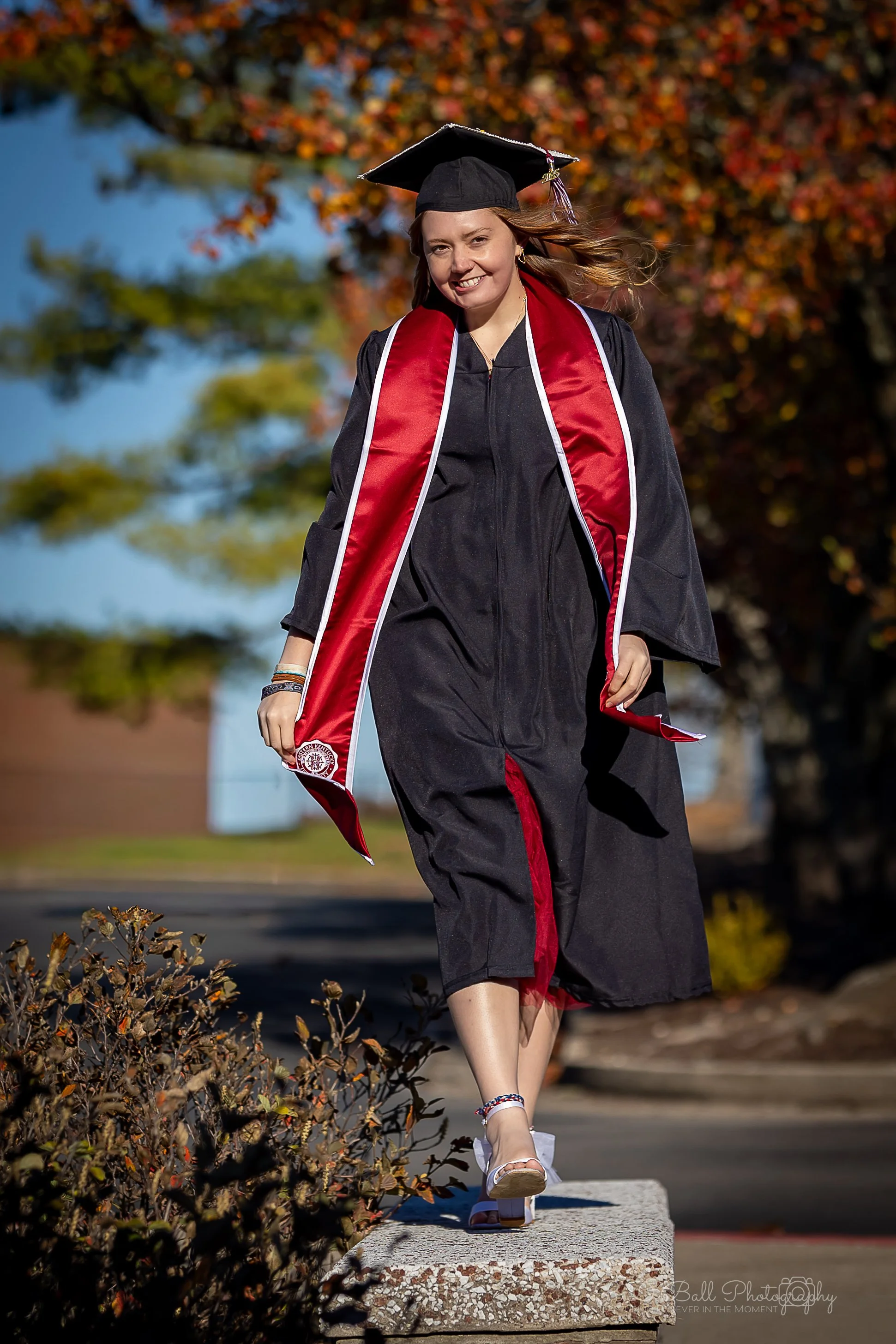A young woman in a graduation cap and gown walking outdoors in the sunlight, with autumn trees in the background.