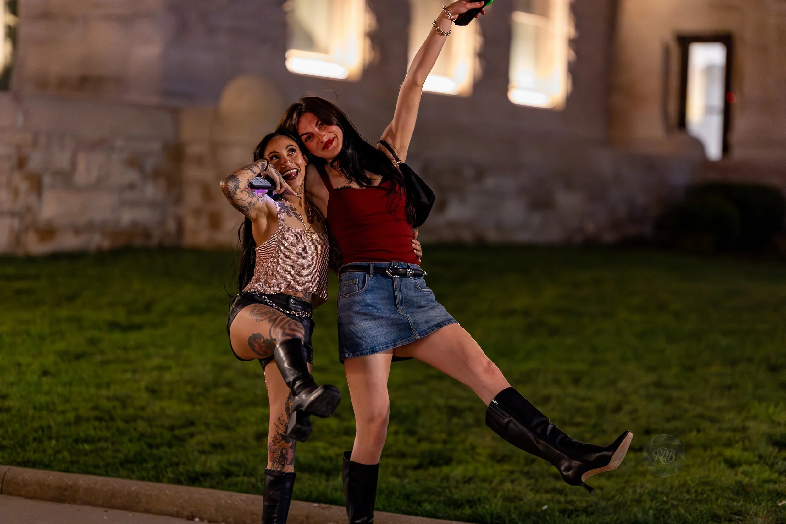 Two women taking a selfie outdoors at night, one with colorful tattoos and wearing a sleeveless top and shorts, the other in a red tank top and denim skirt, both smiling and wearing black boots.