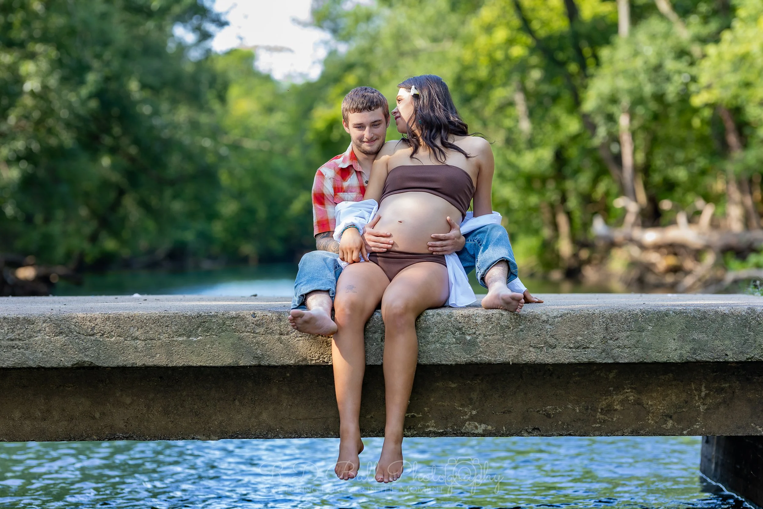 A pregnant woman and a man sitting on a dock by a river, with lush green trees in the background. The man is hugging her from behind and they are smiling at each other. The woman is wearing a brown strapless top and shorts, and the man is wearing a r