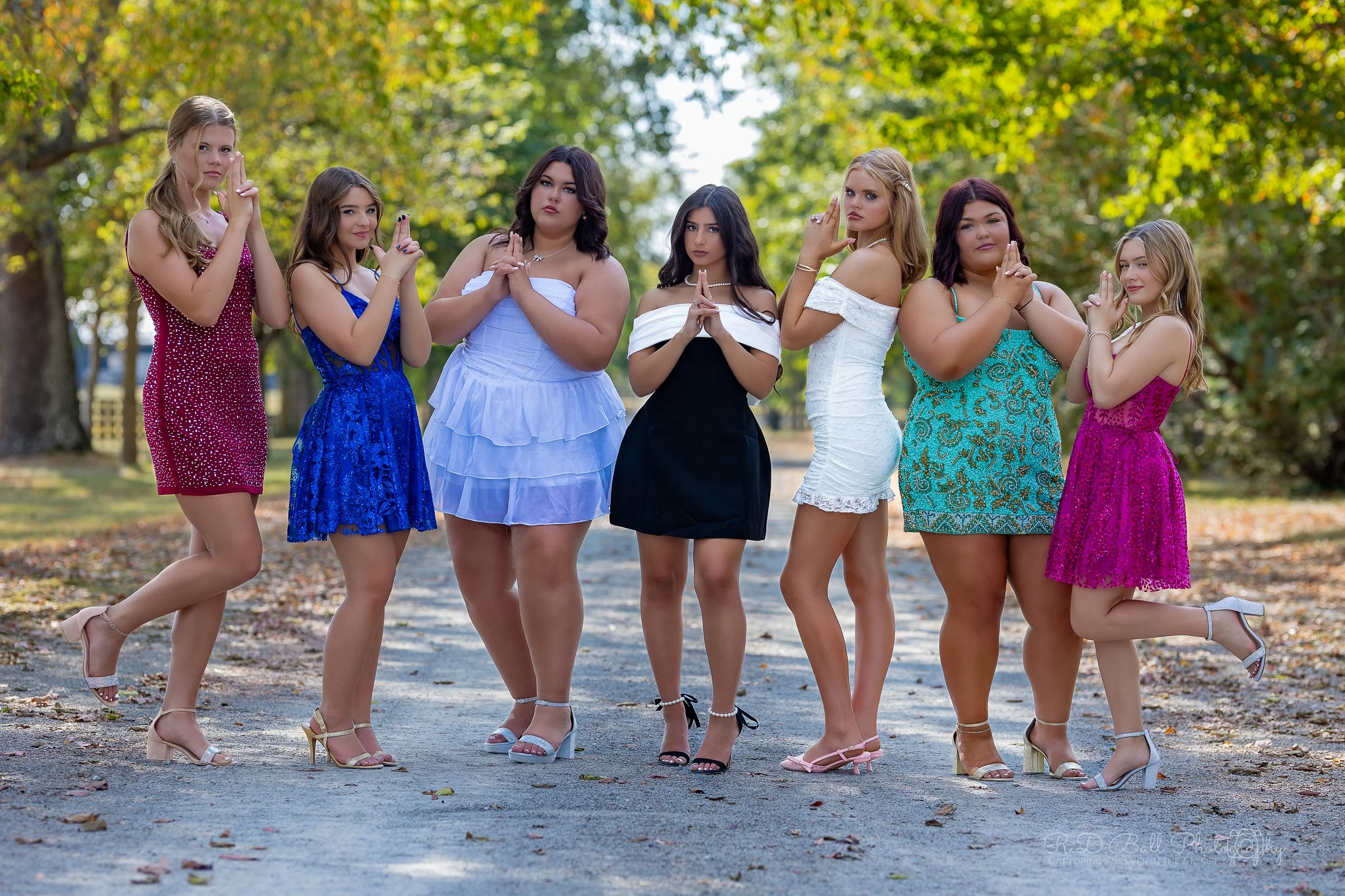 Seven young women standing outdoors on a path surrounded by trees with leaves. They are dressed in colorful dresses and high heels, standing in a line with their hands clasped together near their chests, appearing to pose.