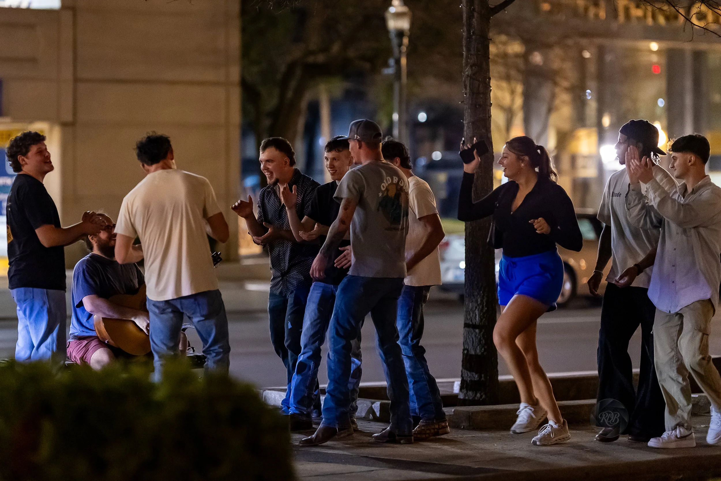 Group of young people dancing and socializing on a sidewalk at night in an urban area, with a guitarist sitting on a chair playing music.