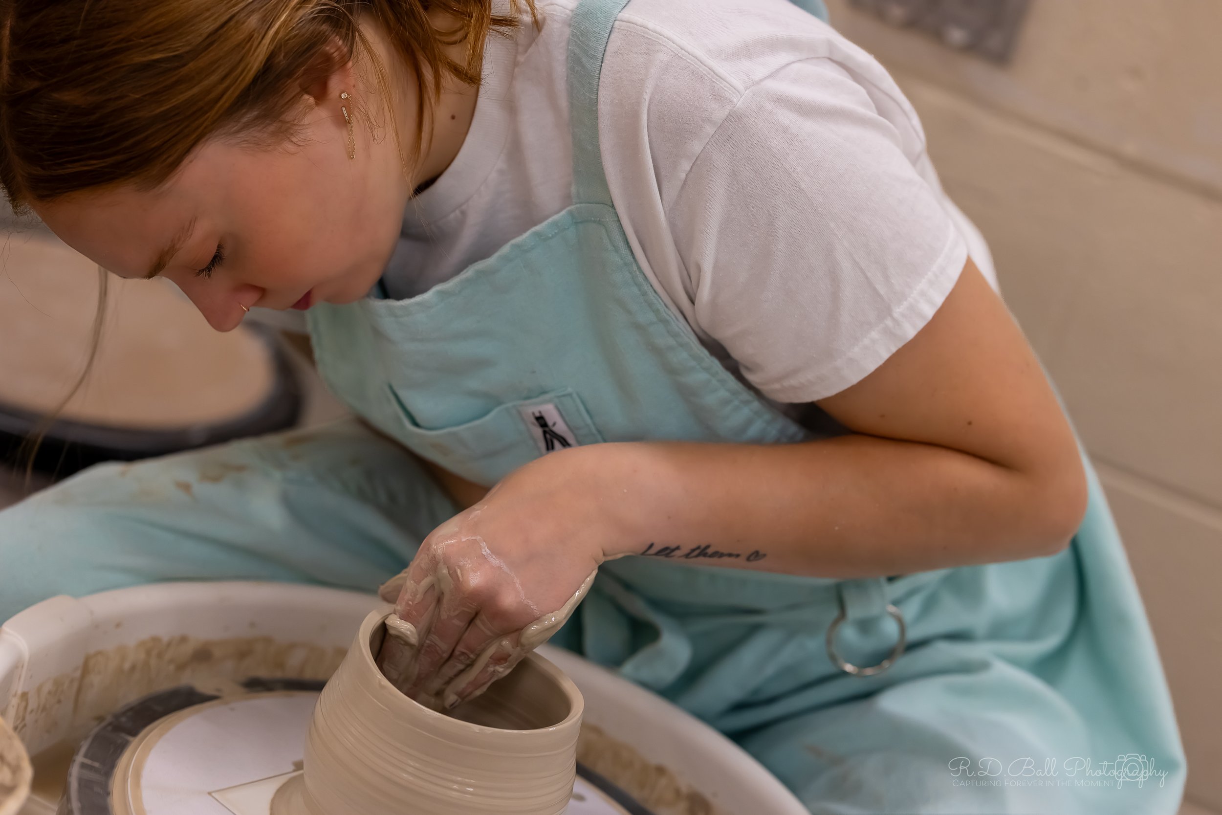 A woman with red hair and earrings crouches over a pottery wheel, shaping clay with her hands in a pottery studio.