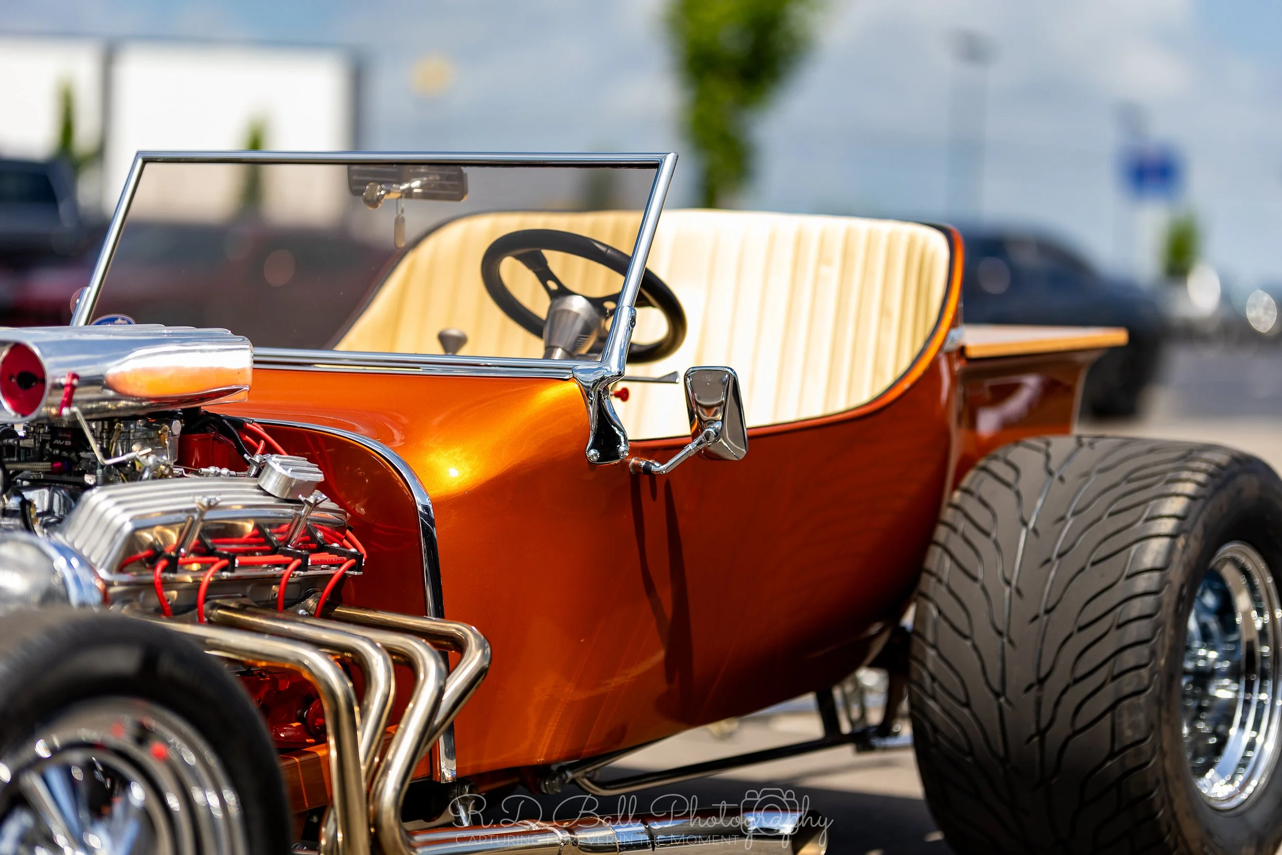 Close-up of a vintage drag racing car with a bright orange body, featuring a visible supercharged engine with chrome pipes, a black steering wheel, and a cream-colored seat. The car is outdoors with a parking lot and blurred background.