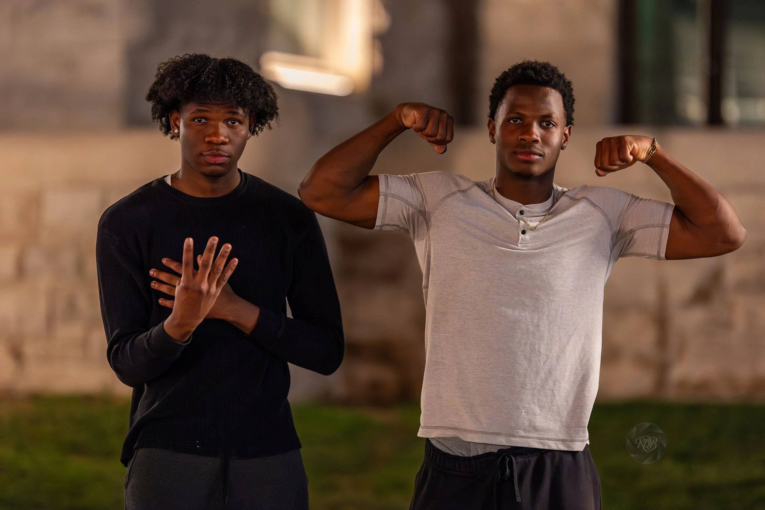 Two young men, one with curly hair wearing a black shirt holding his hand to his chest, the other with short curly hair in a gray t-shirt flexing his arm muscles, standing outdoors with a blurred background.