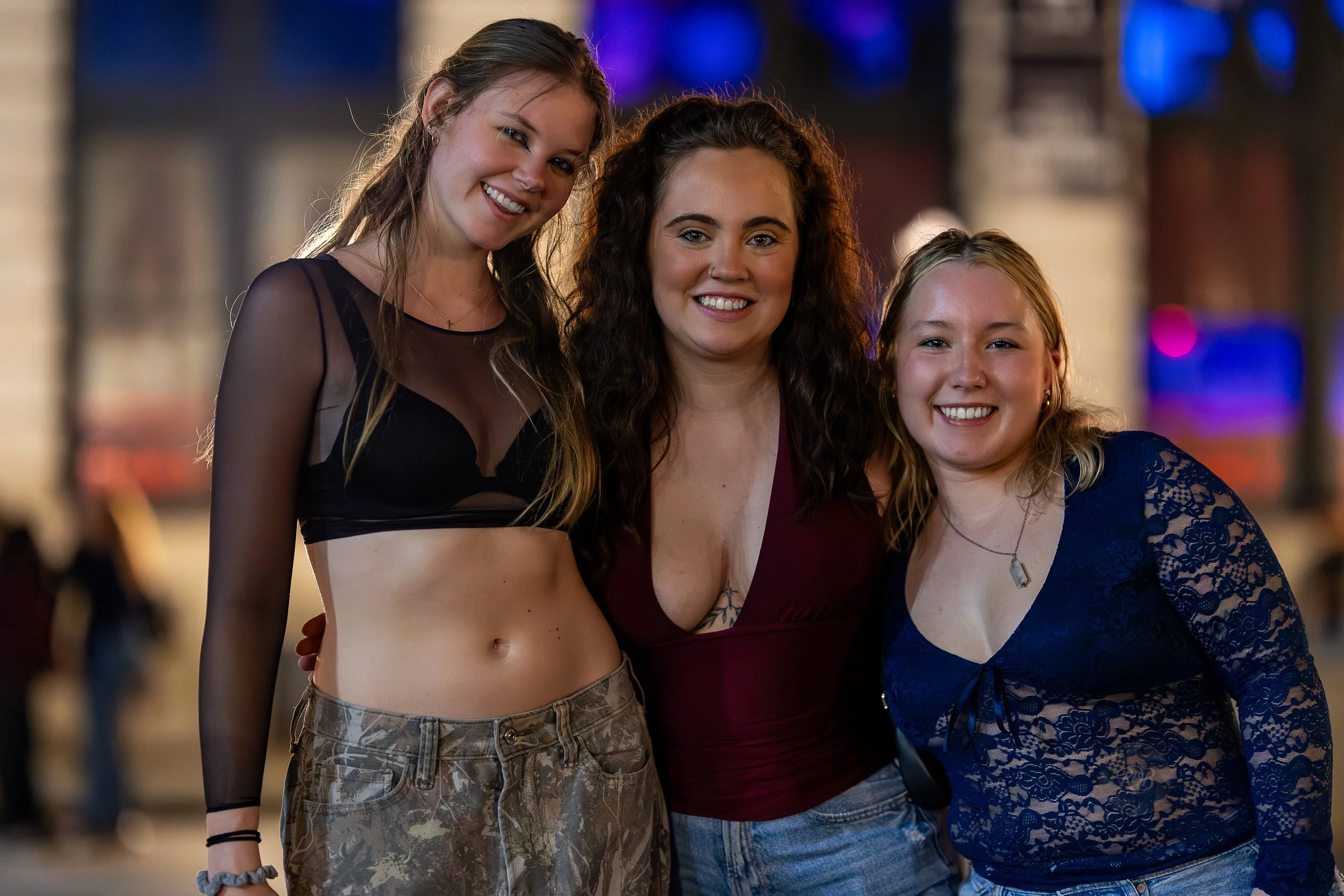 Three women smiling and posing together outdoors at night, with a blurred background of a building and purple and pink lights.