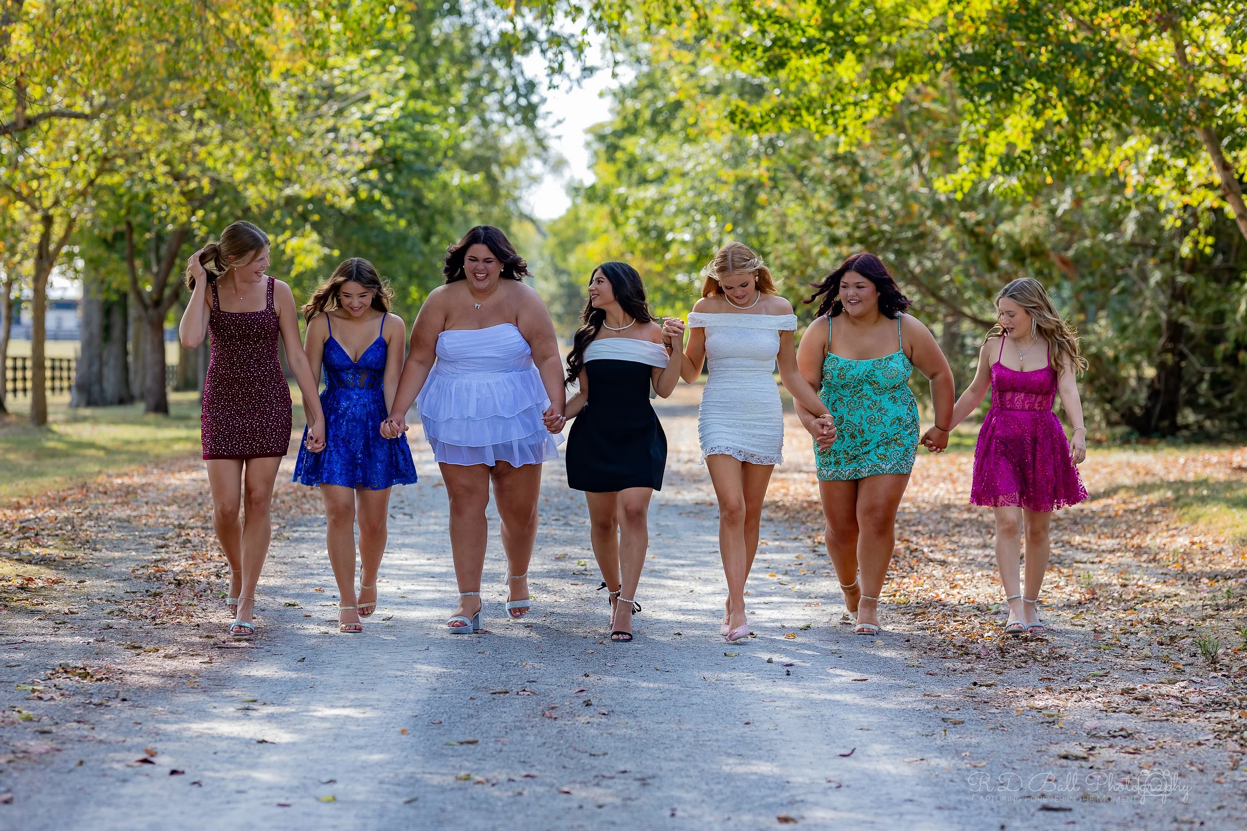 Group of seven women walking hand in hand on a tree-lined path, smiling and enjoying the outdoors.