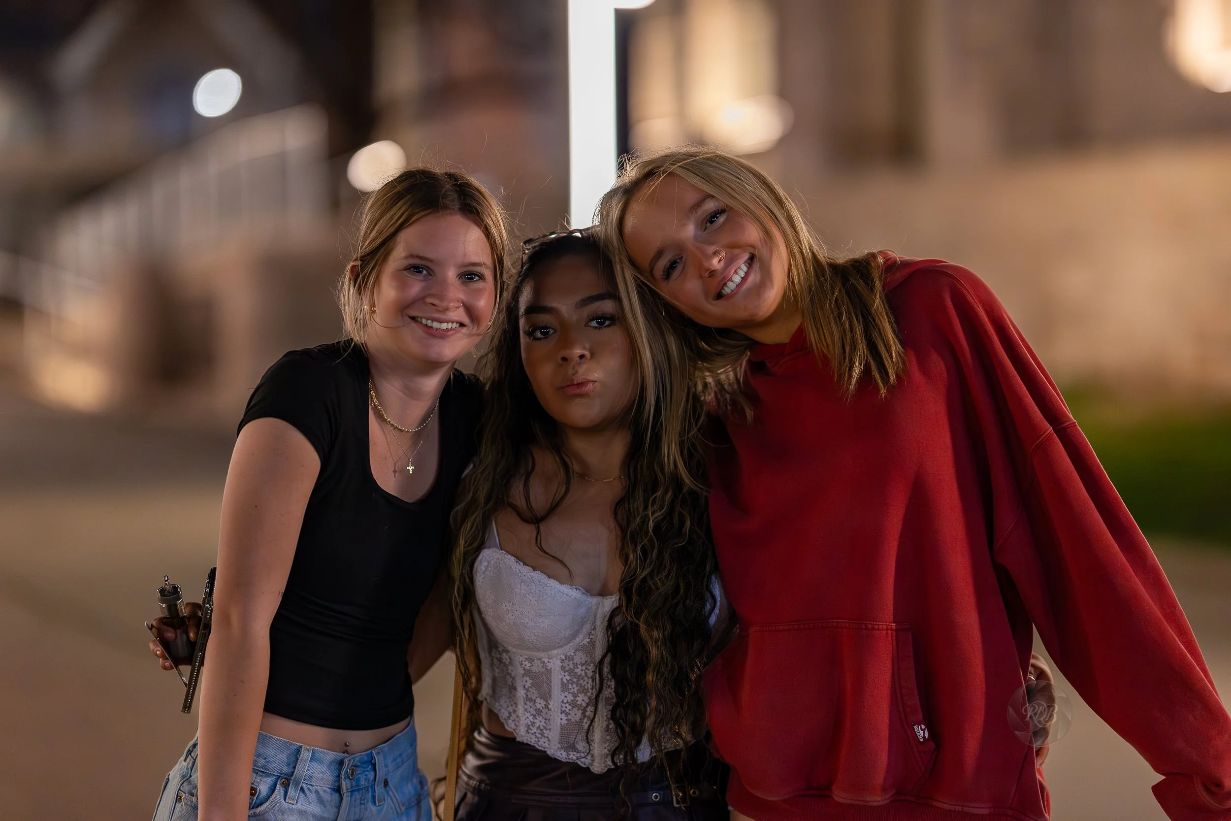 Three young women standing close together on a city sidewalk at night, smiling at the camera.