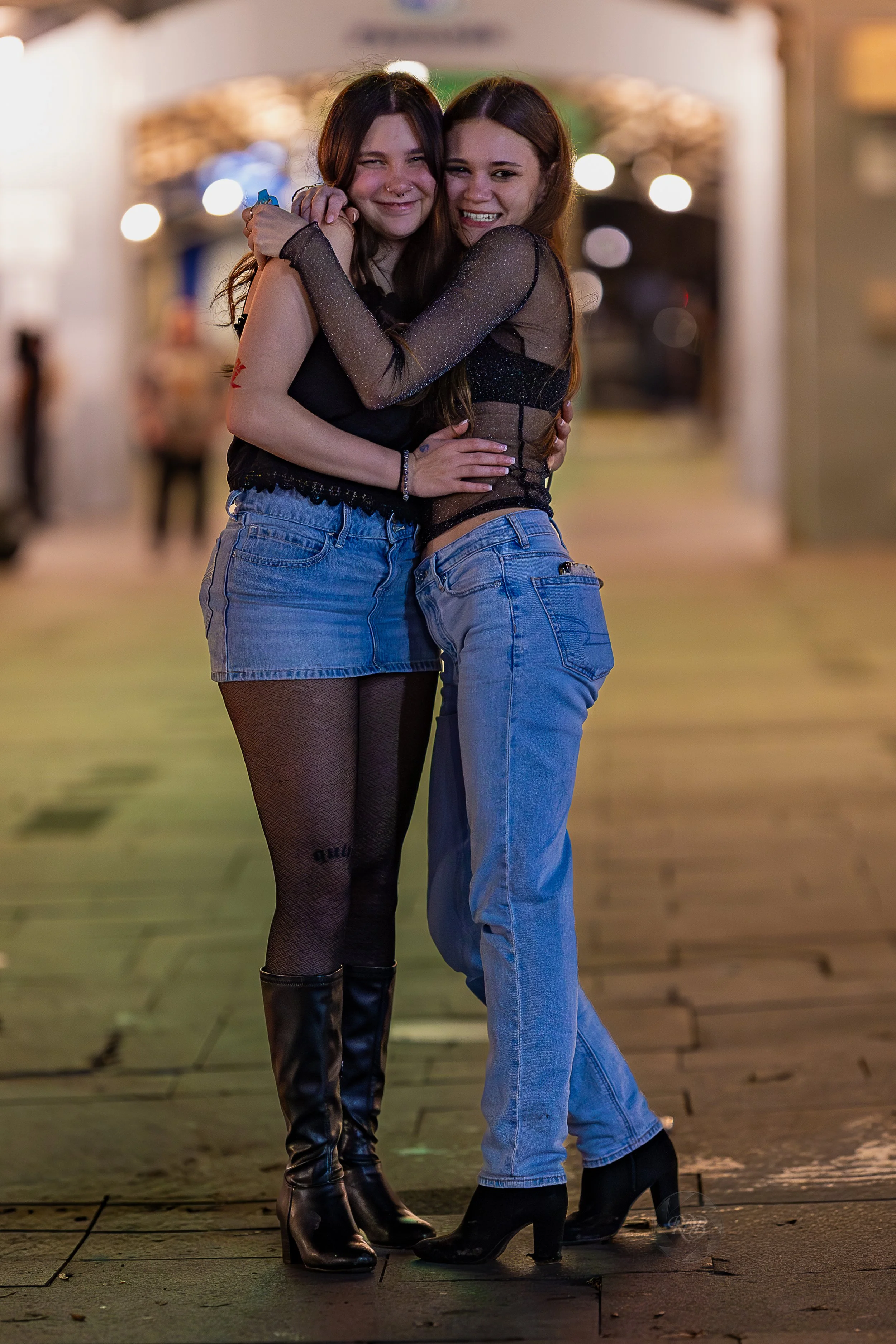 Two young women hugging and smiling on a city street at night, illuminated by streetlights and illuminated signs.