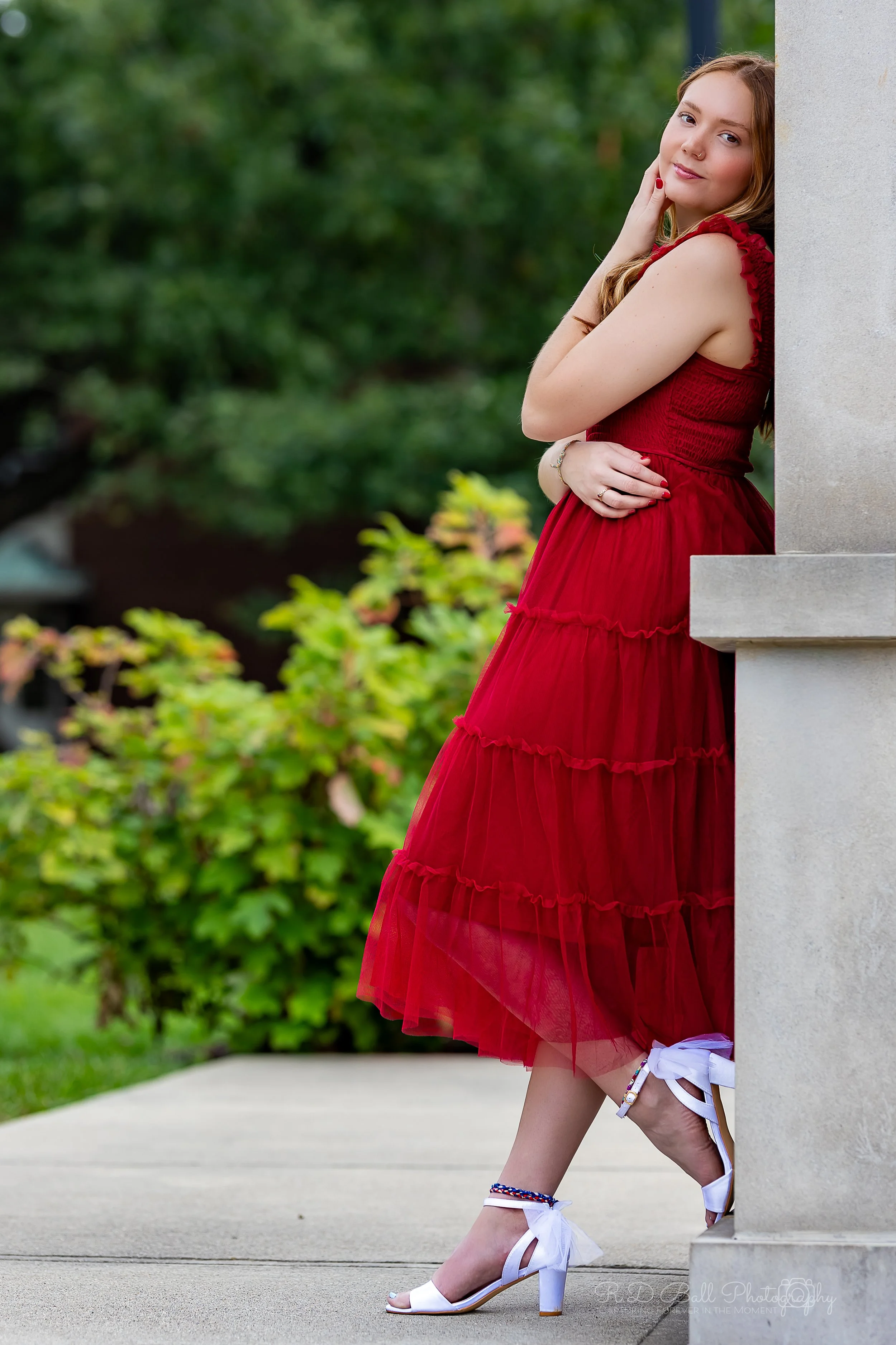 Young woman in a red dress leaning against a stone structure outdoors with greenery in the background.