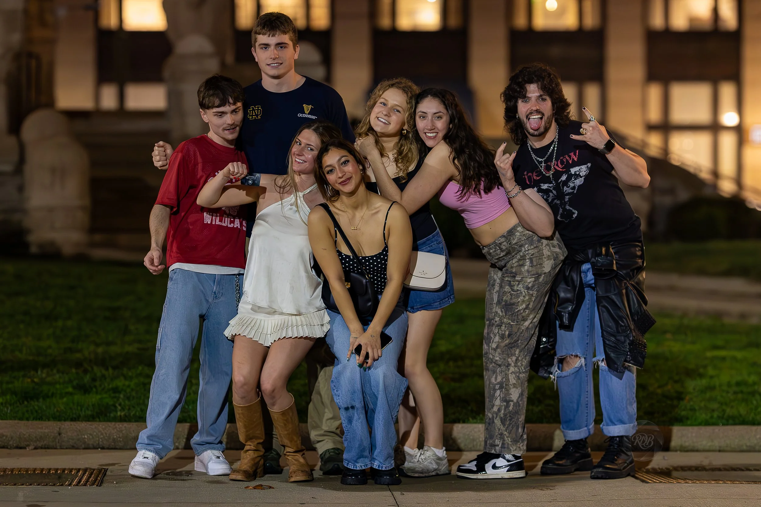 Group of nine young friends posing together outdoors at night, smiling and making playful gestures.