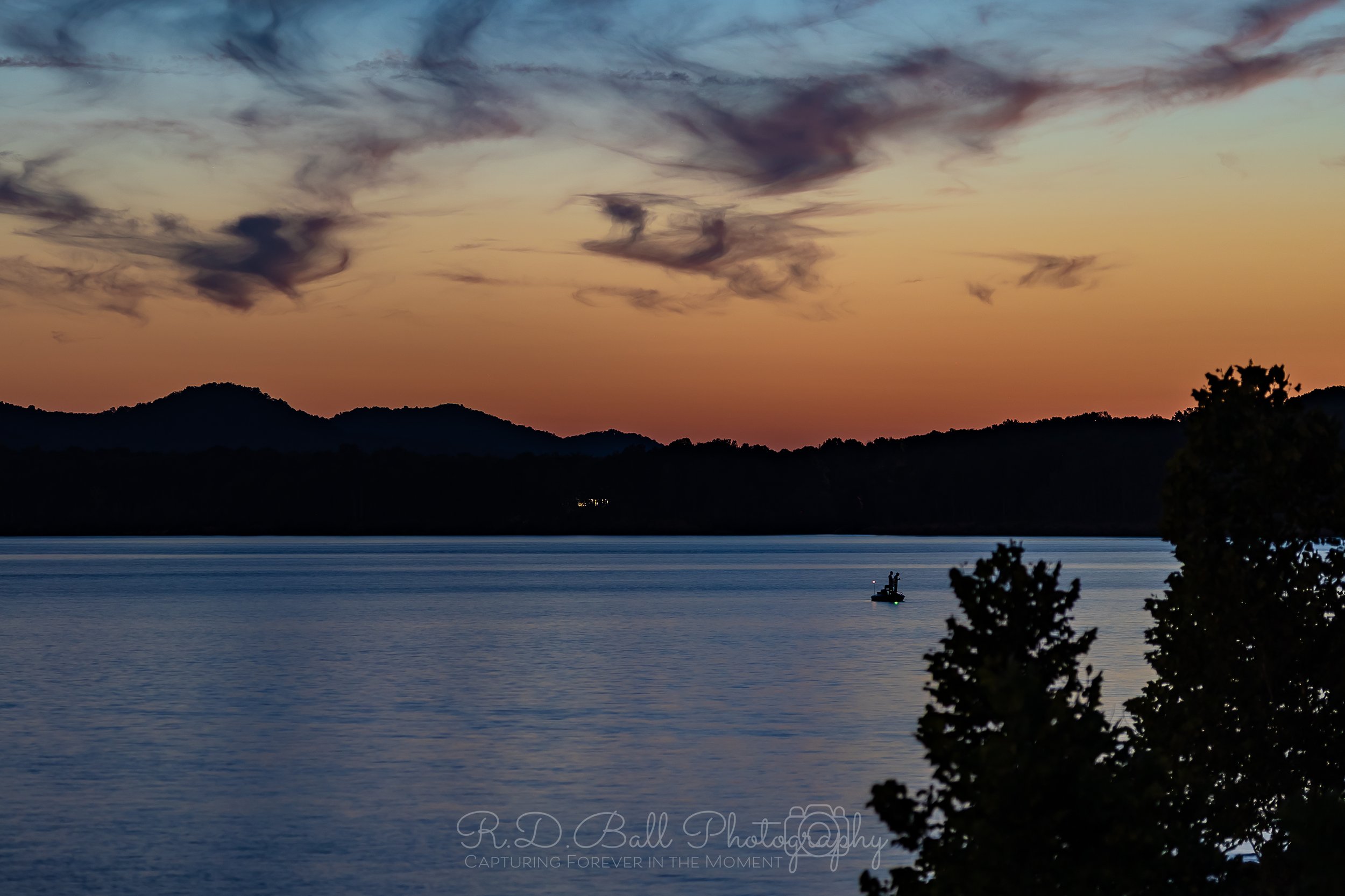Sunset over a lake with hills in the background and a small boat with two people on the water. Silhouetted trees in the foreground.