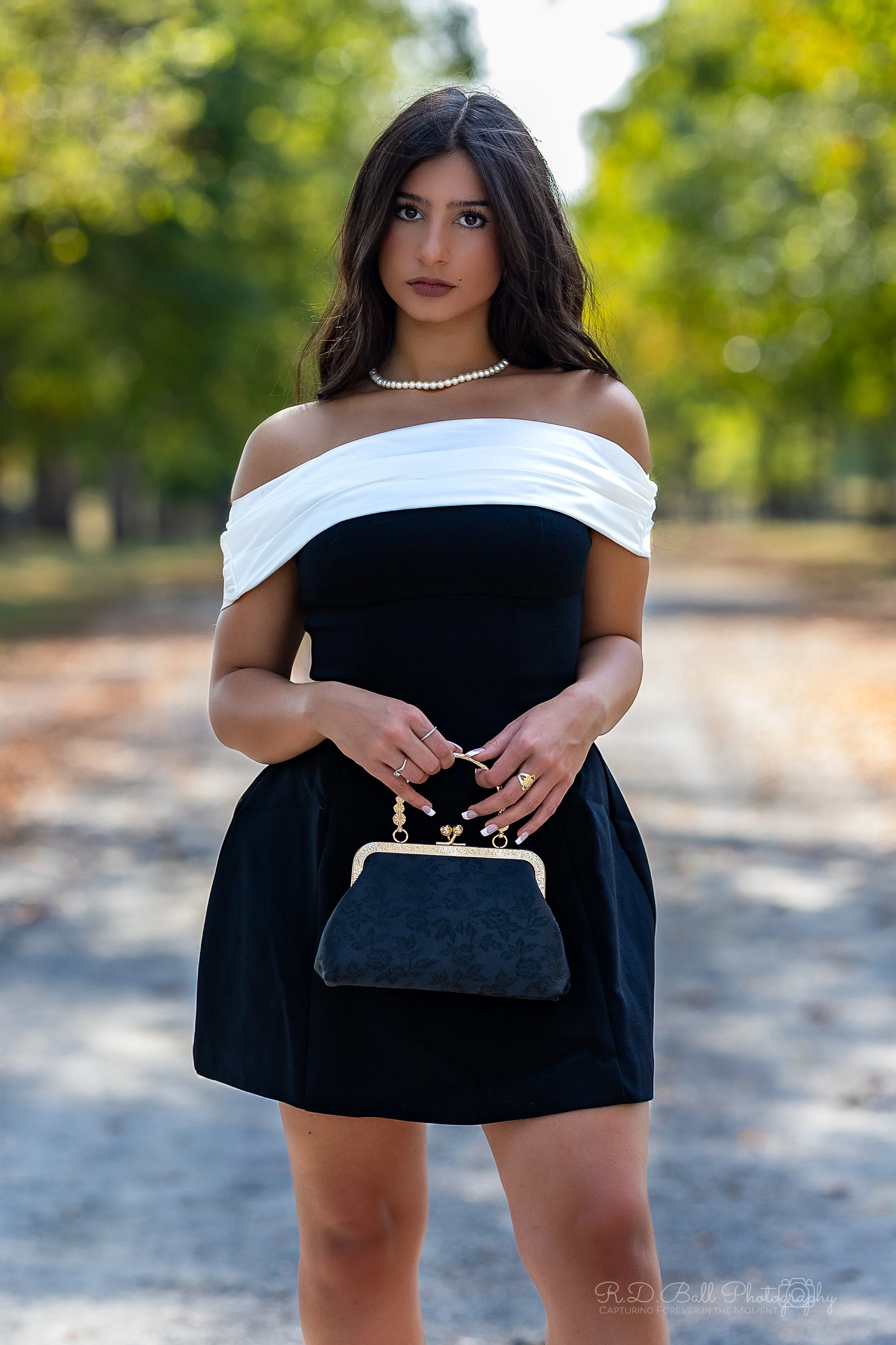 Young woman standing outdoors holding a black purse, dressed in a black and white off-the-shoulder dress, wearing a pearl necklace, with a blurred green and yellow background.