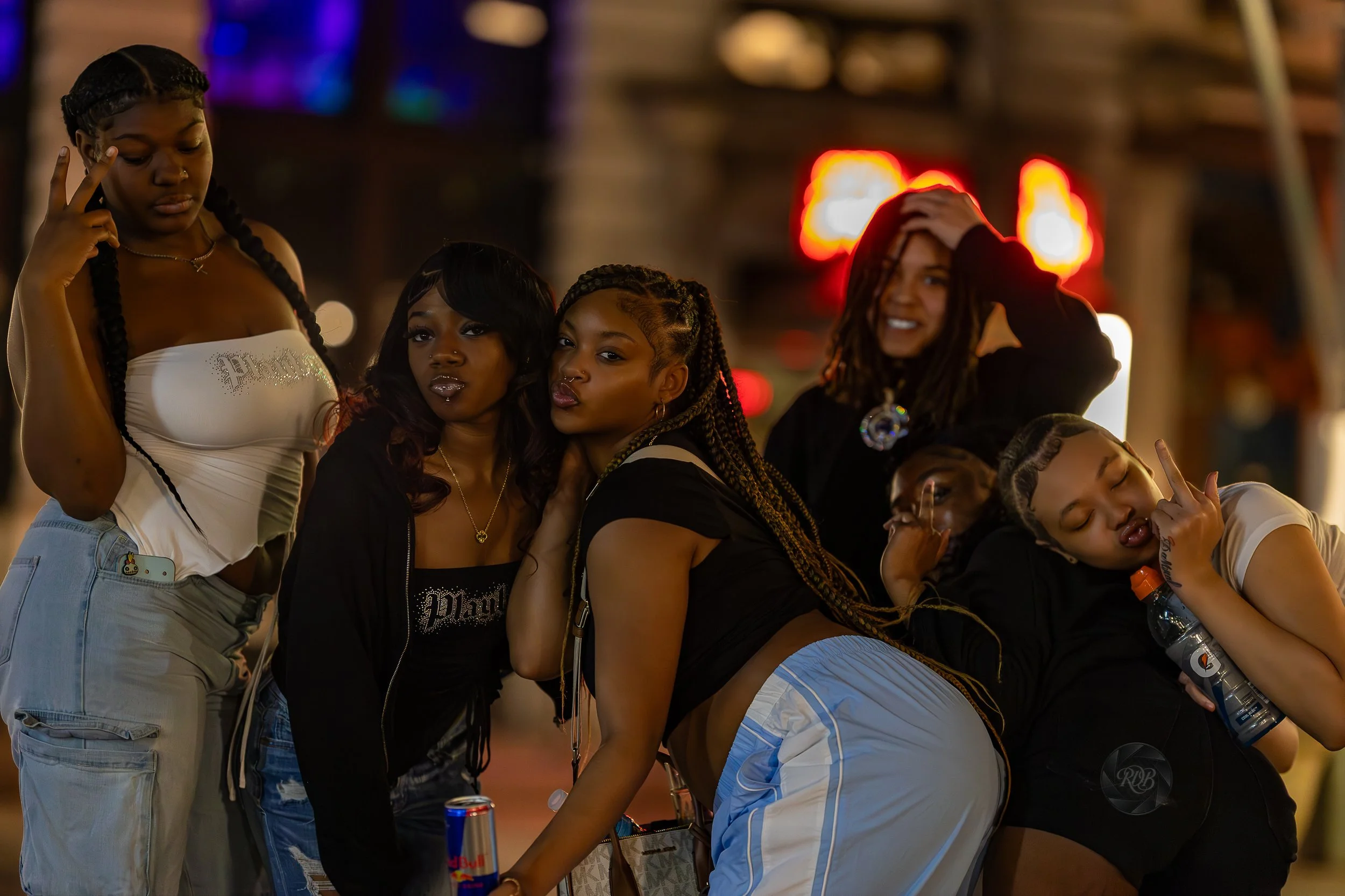 Group of six young women with braided hair posing on a city street at night, some making playful gestures, in casual trendy clothing with neon city lights in the background.
