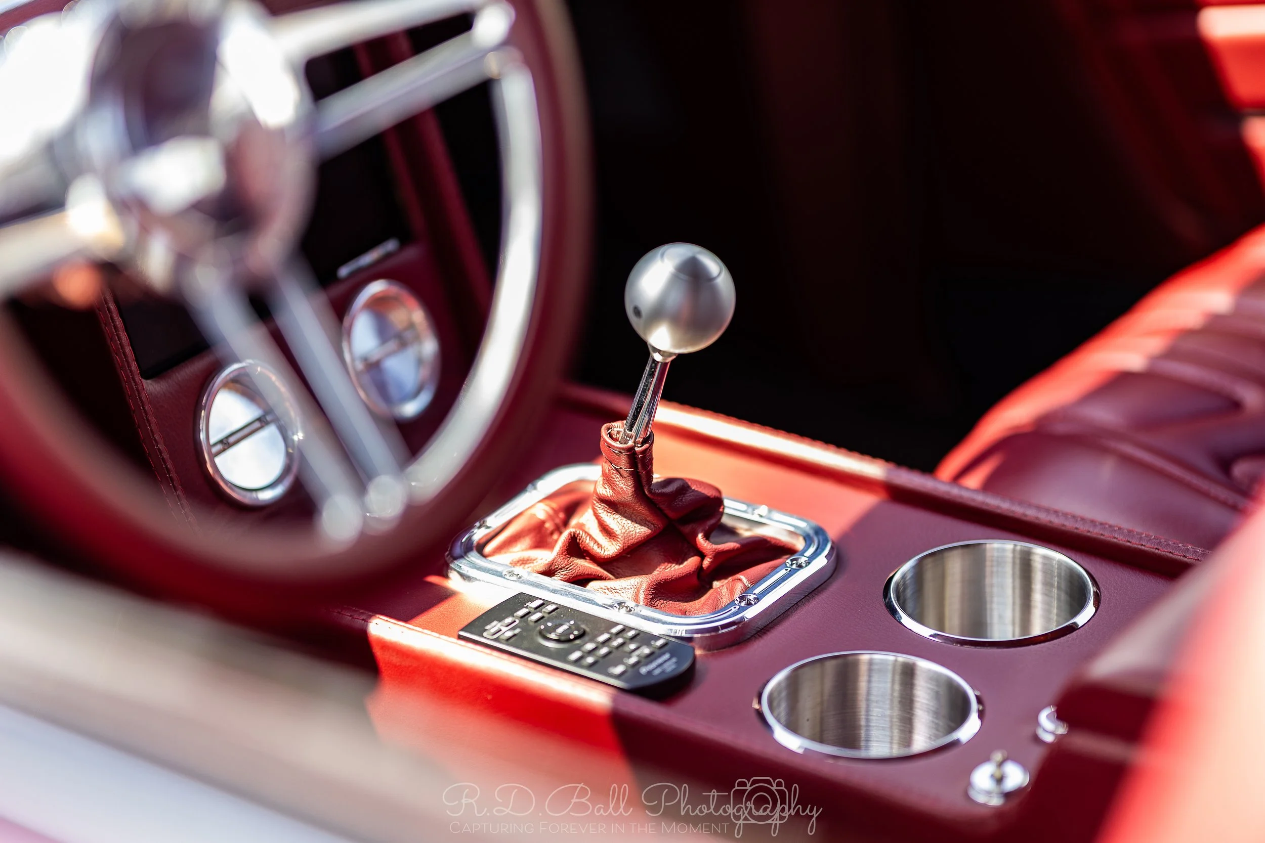 Close-up of a vintage car's interior featuring a manual gear shift with a metallic ball shifter, red leather upholstery, a dashboard with gauges, and a remote control placed on the center console.