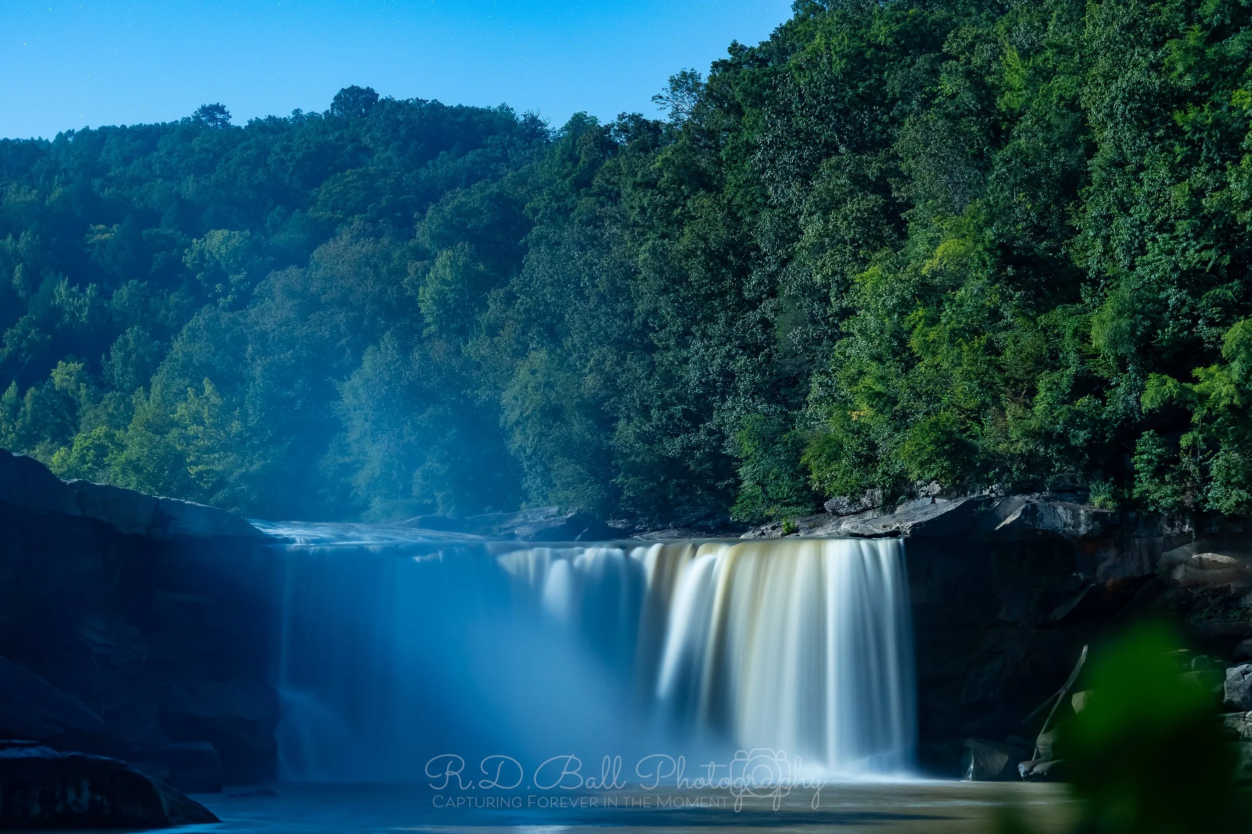 A waterfall flowing over rocks into a river, surrounded by lush green trees and mist rising from the water.