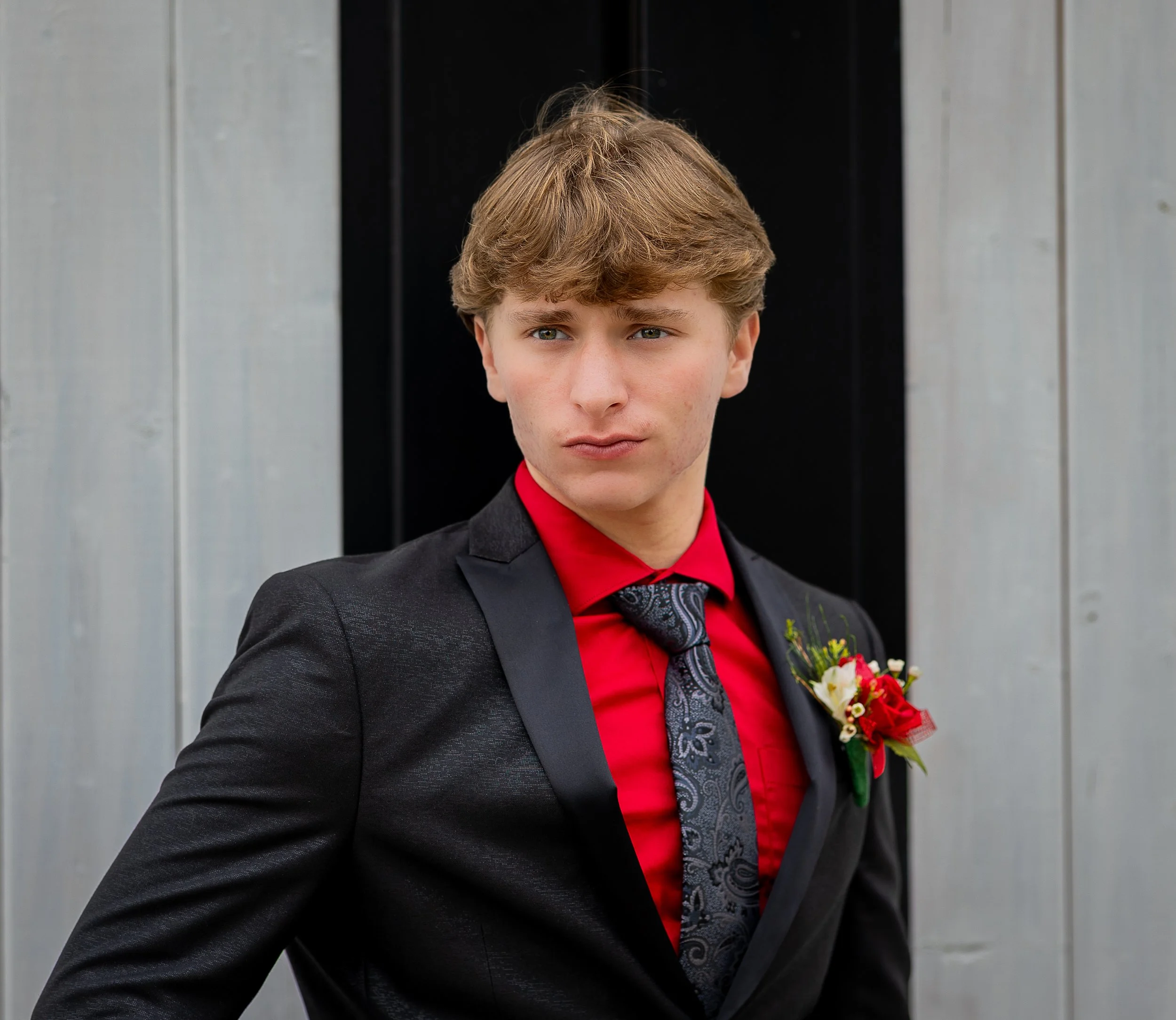 A young man with blond hair and blue eyes dressed in a black suit, red shirt, and black patterned tie, standing in front of a gray and black background with a flower boutonniere on his lapel.