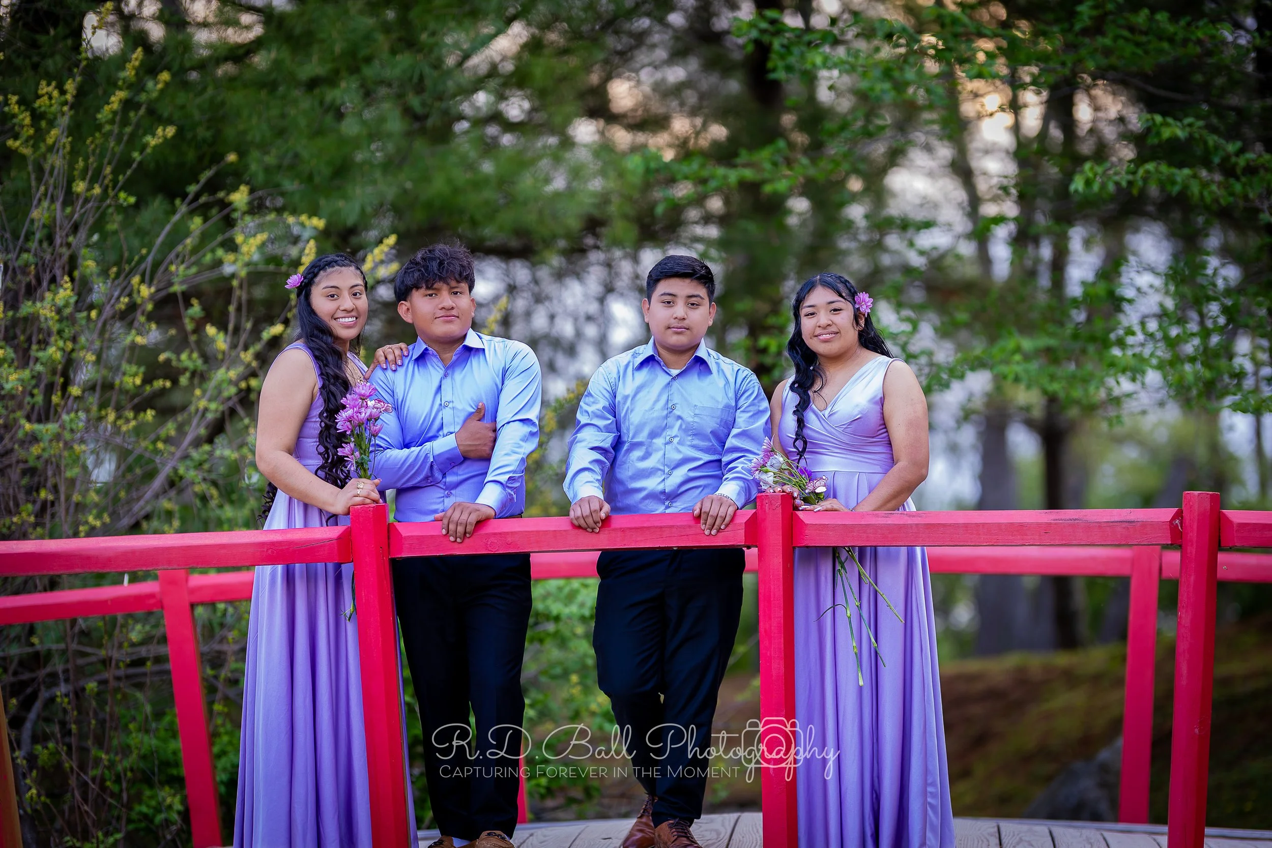 Group of four teenagers standing on a red wooden bridge outdoors, two girls in lavender dresses holding flowers, and two boys in blue shirts with one giving a thumbs-up, surrounded by trees.